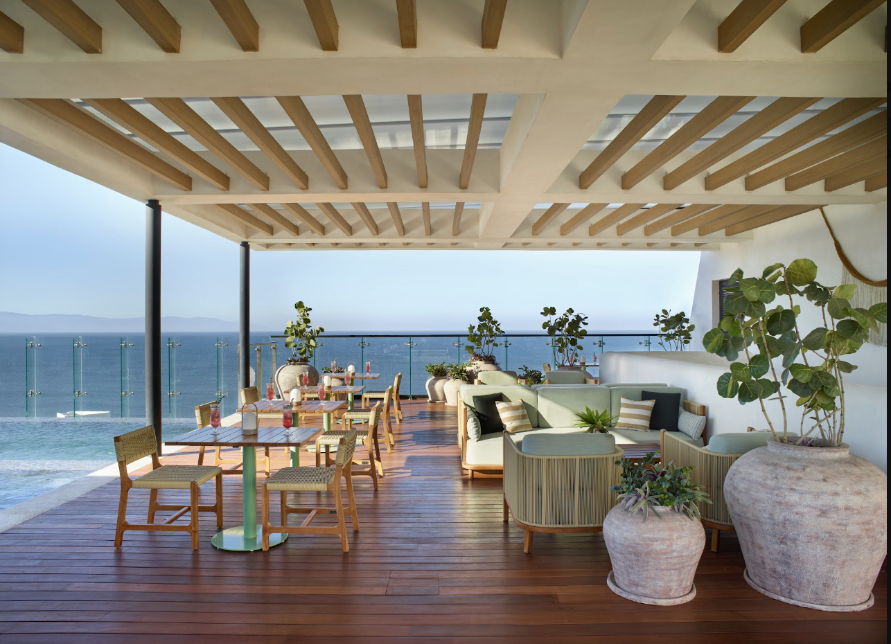 altorooftop2 be all inclusive Open-air seaside terrace with a wooden deck, glass railing, and tables with chairs, overlooking the ocean. Cushioned seating and potted plants along the edge.
