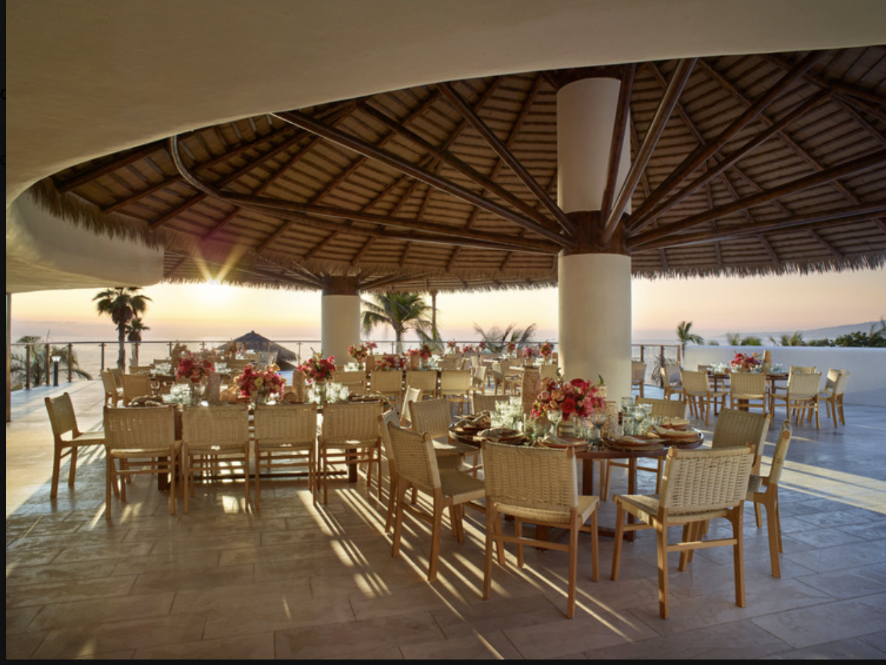 aquamadreterr1 be all inclusive Beachfront outdoor dining setup under a thatched pavilion at sunset, with round tables, wooden chairs, and pink flower centerpieces by the sea.