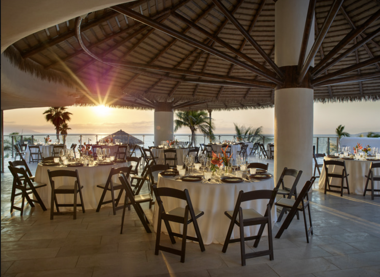 aquamadreterr2 be all inclusive Open-air beachside restaurant at sunset with round tables, white tablecloths, and wooden chairs under a thatched roof.