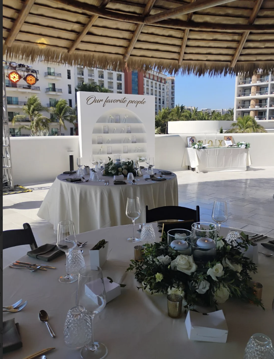 aquamadreterr3 be all inclusive Outdoor rooftop reception with round tables, white linens, floral centerpiece, and candle decor under a thatched roof.