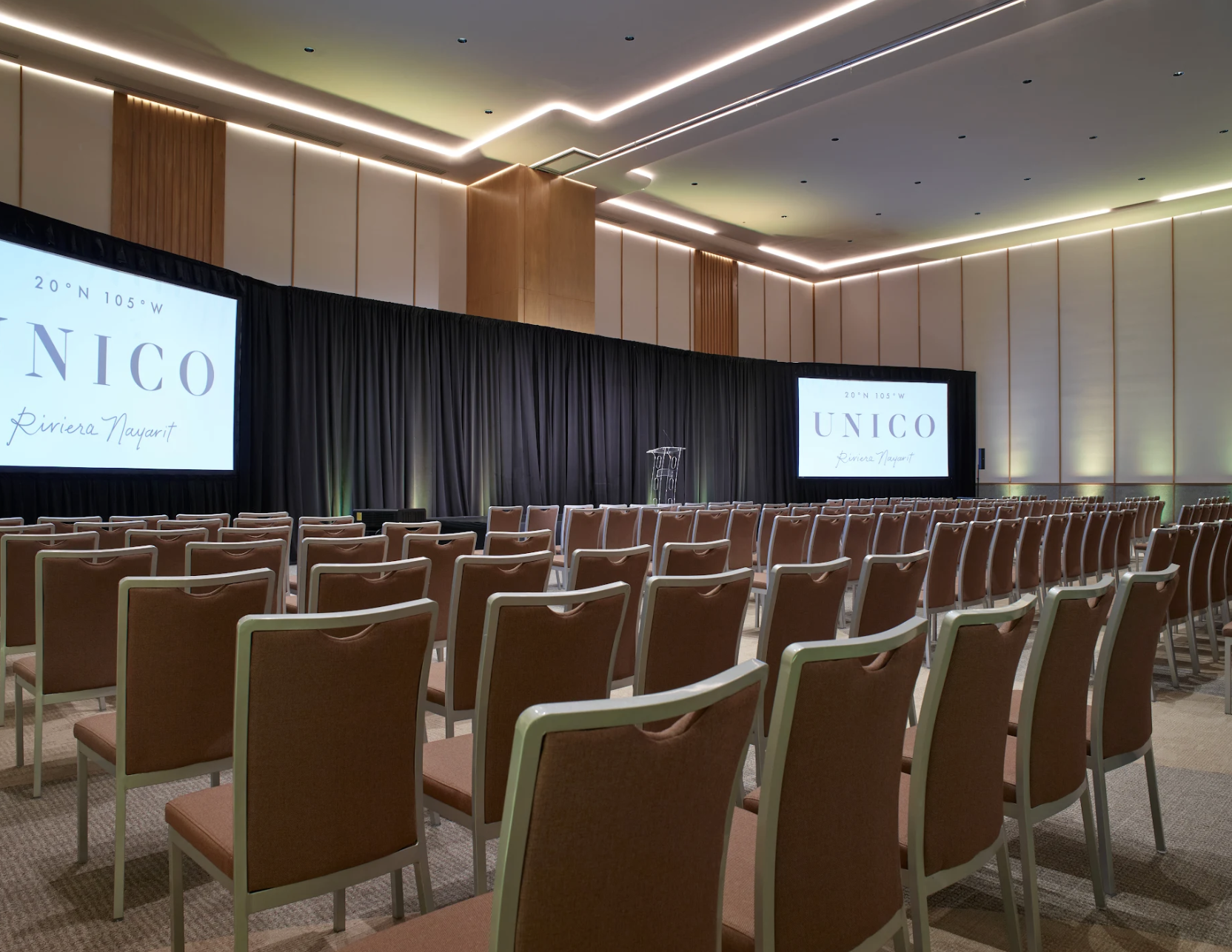 ballroomconf2 be all inclusive Conference hall set up for a presentation with a stage, black curtain backdrop, and two projection screens displaying a logo, facing rows of brown chairs.