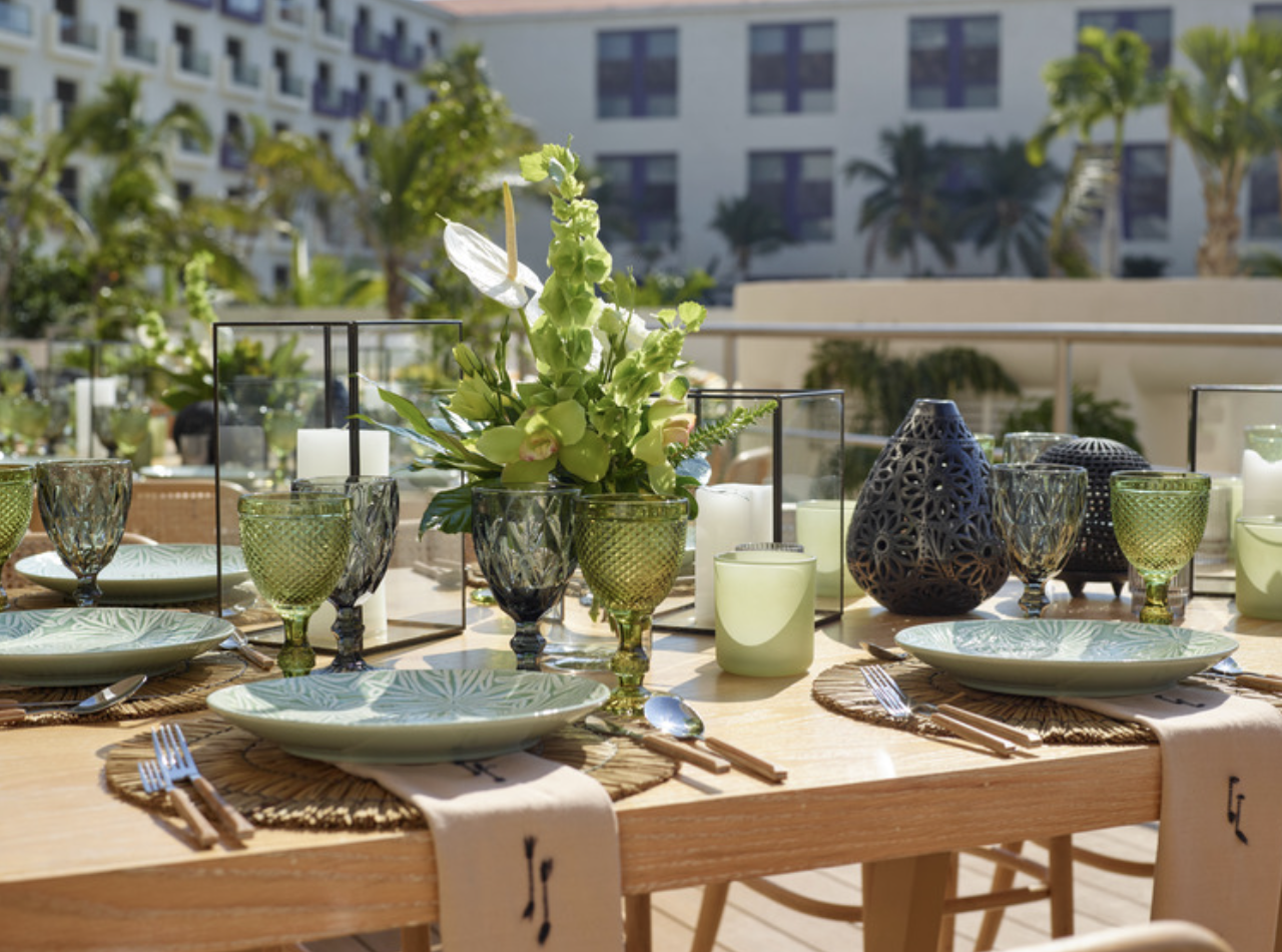 inezterr3 be all inclusive Outdoor dining table with green glassware, plates, and a floral centerpiece on a sunlit balcony.
