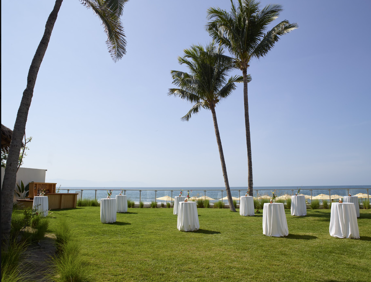 pacificgarden1 be all inclusive Outdoor beachfront event setup with white-draped cocktail tables on a green lawn and palm trees overlooking the ocean.