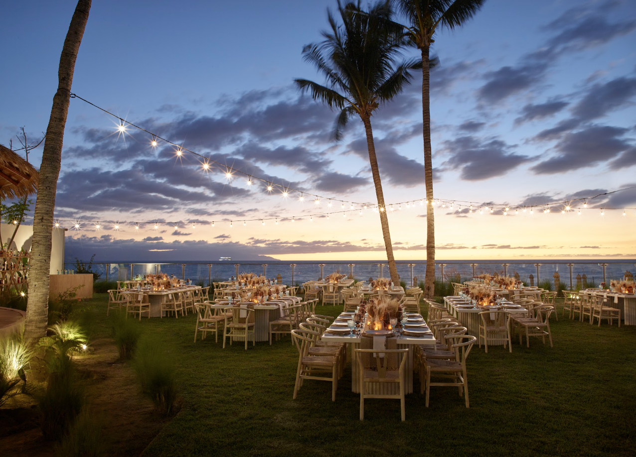 pacificgarden2 be all inclusive Beachfront outdoor dinner setup at dusk with string lights between palm trees and ocean in the background.