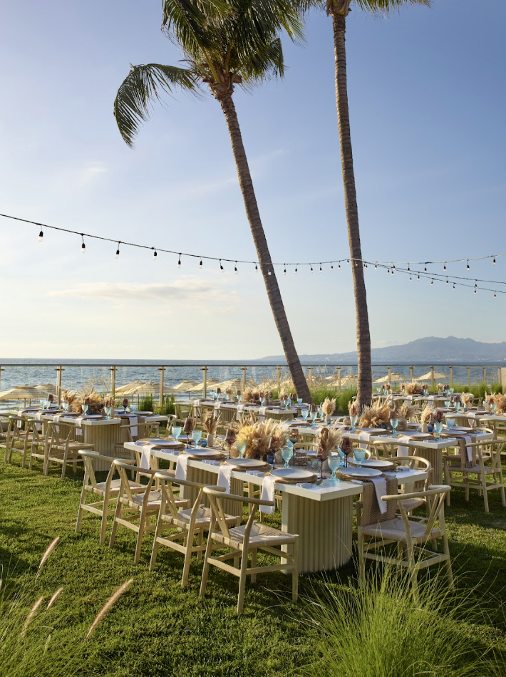 pacificgarden3 be all inclusive Outdoor beachside banquet on a grassy lawn with round tables, white linens, blue glassware, and string lights between palm trees by the water.",