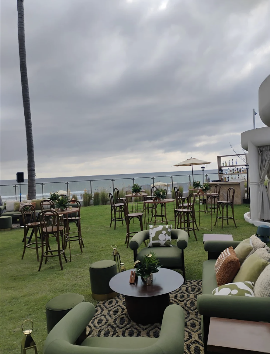 pacificgarden4 be all inclusive Outdoor lounge area with green sofas, round tables, and a bar by the sea under cloudy skies