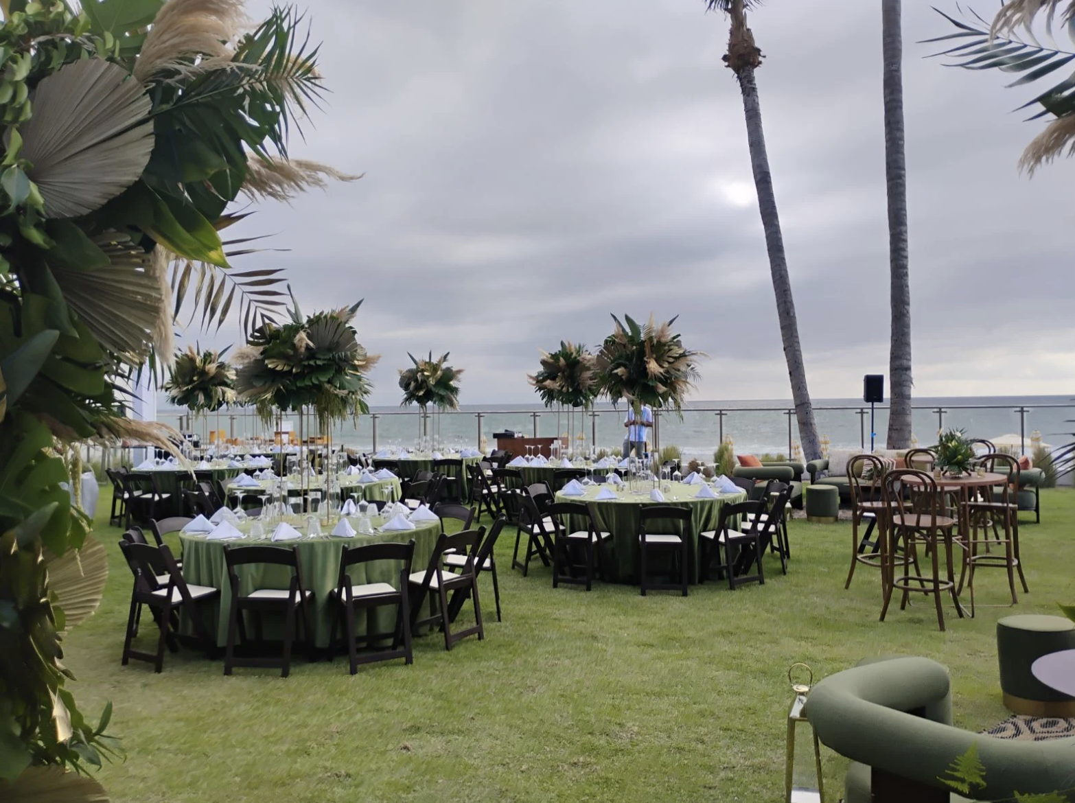 pacificgarden5 be all inclusive Outdoor seaside banquet setup with round tables, green tablecloths, white napkins, and dark chairs on a grassy lawn with palm trees and the ocean in the background, under a cloudy sky.