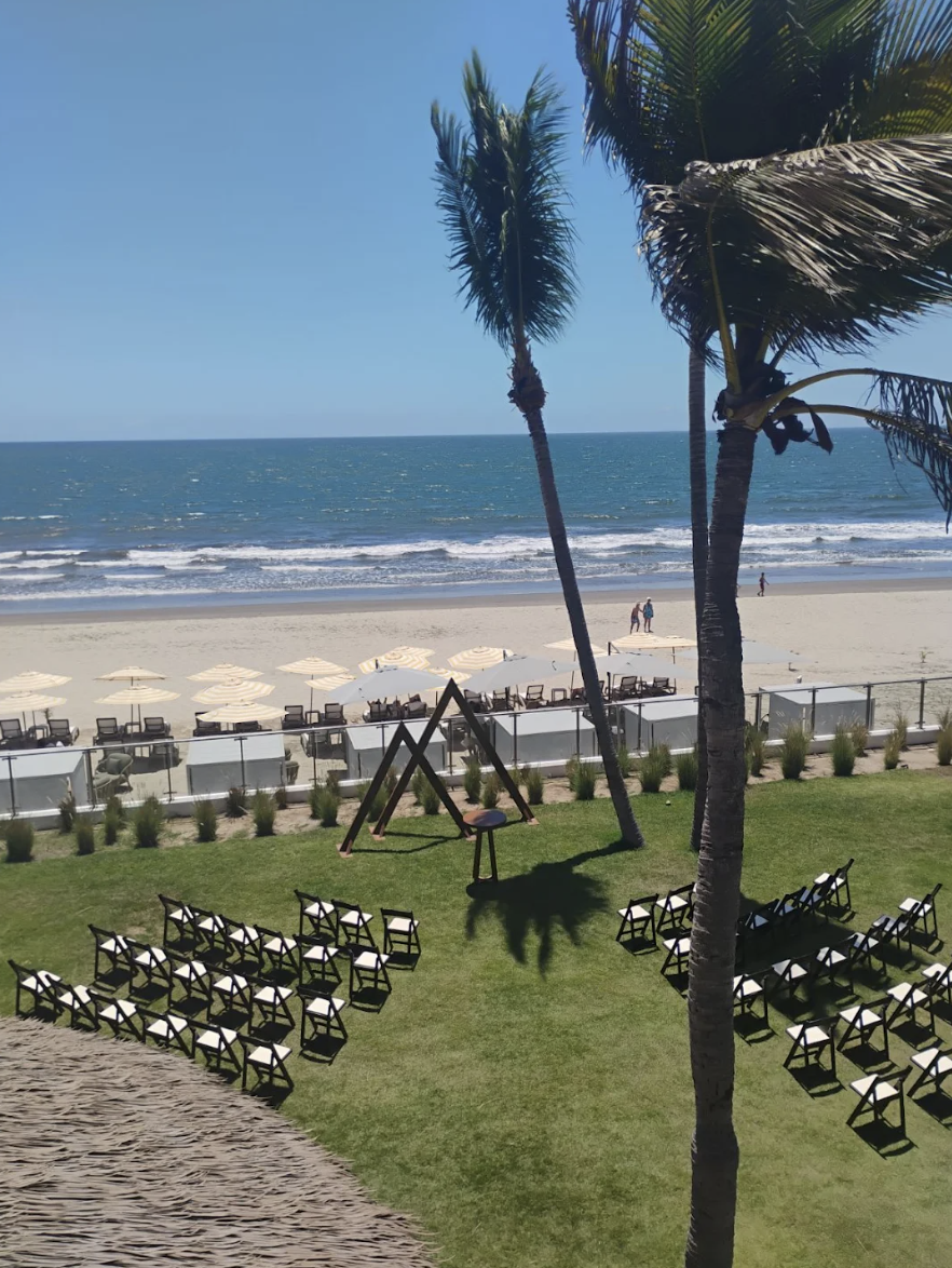 pacificgarden6 be all inclusive Beachside event setup with rows of black chairs on a green lawn facing a small triangular stage; sandy beach, umbrellas, and the ocean in the background.