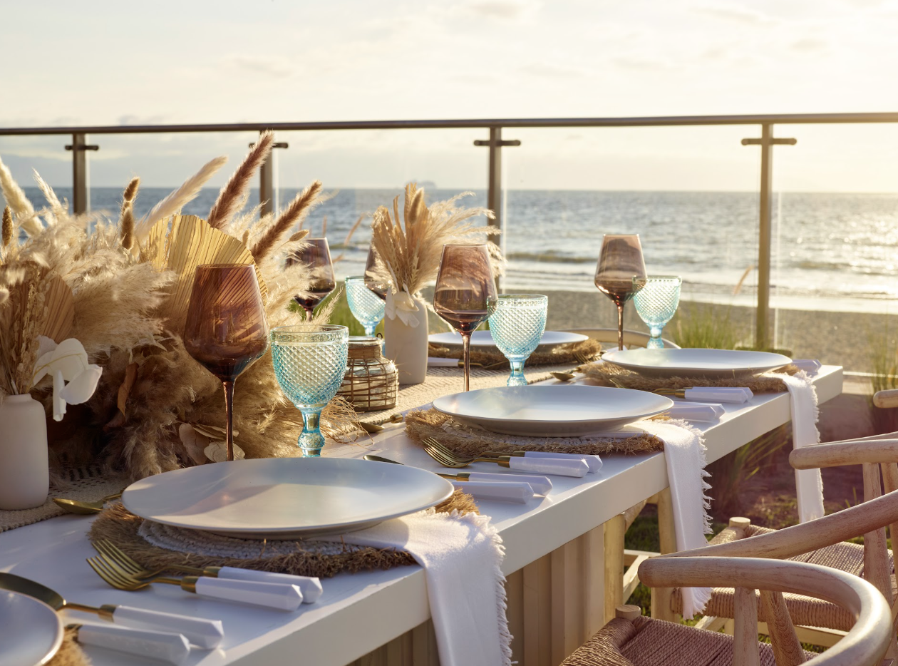 pacificgarden7 be all inclusive Beachside dinner table set for a sunset meal with plates, gold cutlery, and patterned glasses by the ocean