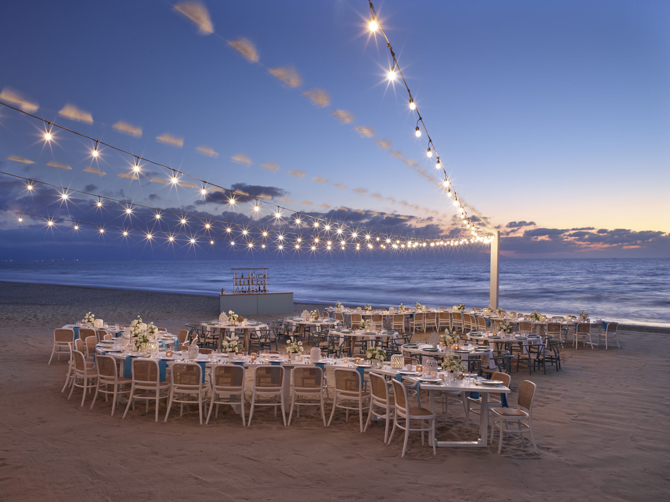 beach vendor set up 2 be all inclusive Beachside evening banquet setup with rows of white tables, chairs, and floral centerpieces under string lights at dusk by the ocean.