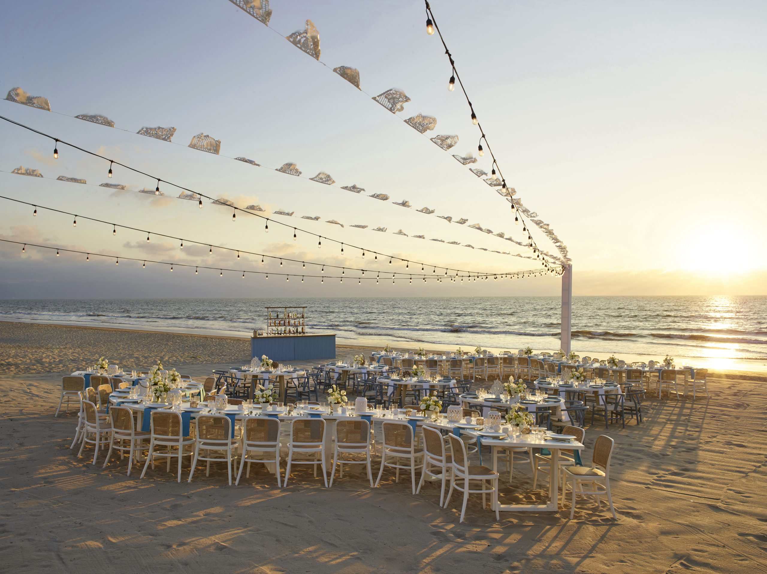 beach vendor set up be all inclusive Rows of white tables and chairs arranged on a sandy beach for an outdoor event, with string lights overhead at sunset and the sea in the background.