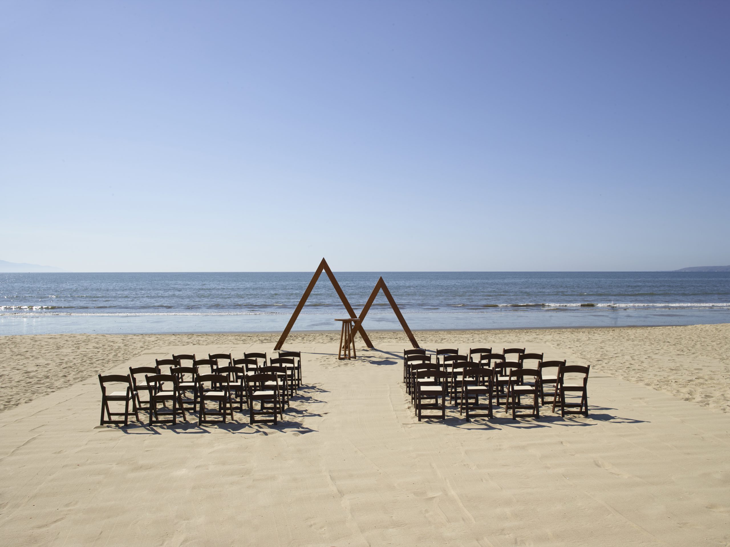 beach wedding be all inclusive Rows of black chairs arranged in a seating area on a sandy beach, facing the calm ocean with a wooden triangular arch centerpiece at the shore.
