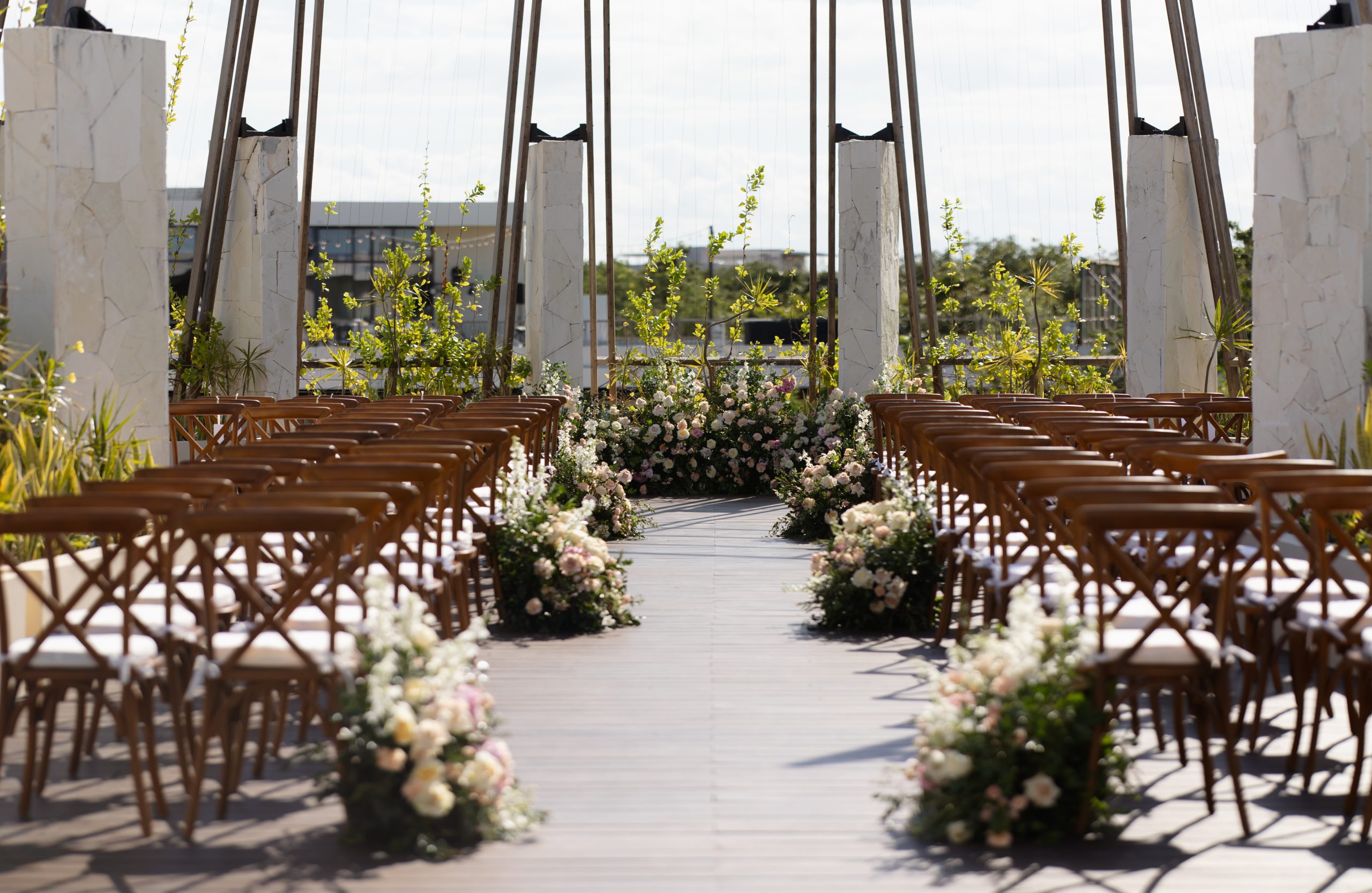 brandon olivia be all inclusive Outdoor wedding aisle with wooden chairs and flower arrangements along a central walkway under stone columns and a wooden framework.