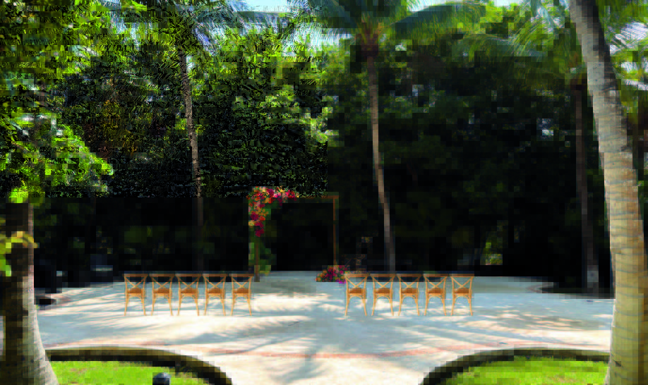 1 be all inclusive Outdoor tropical wedding setup with wooden chairs arranged in two rows facing a flower-decorated arch under palm trees in a sunny garden.