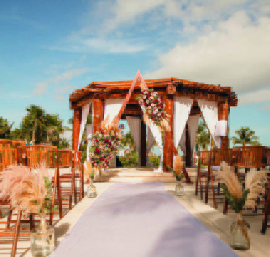 1 be all inclusive Outdoor beach wedding ceremony: wooden arch with flowers and white fabric, lavender aisle runner, and rows of wooden chairs nearby on a sunny day.