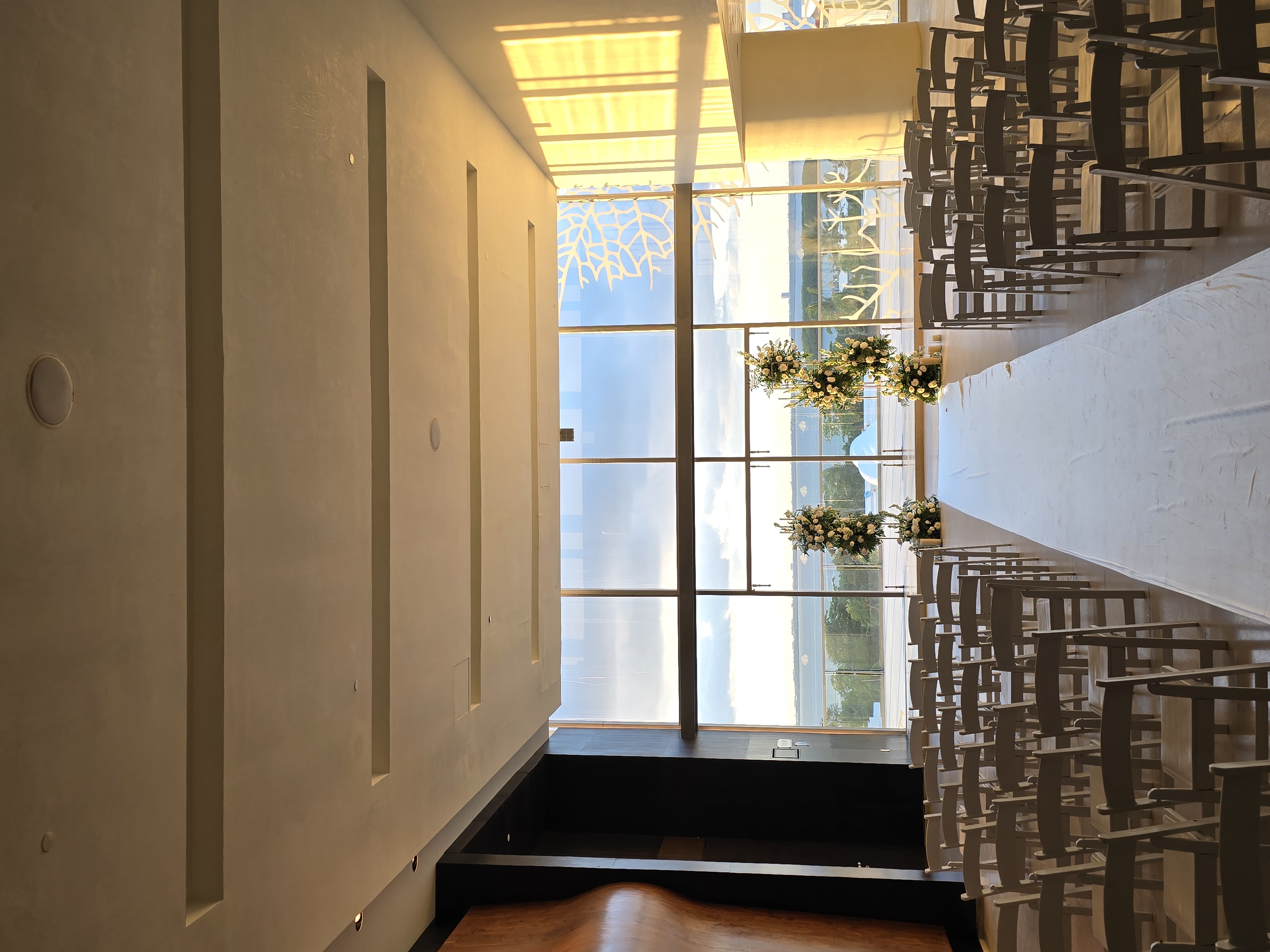 20241206174248 be all inclusive Indoor wedding ceremony space with a long white aisle and multiple rows of chairs facing large windows overlooking the water.