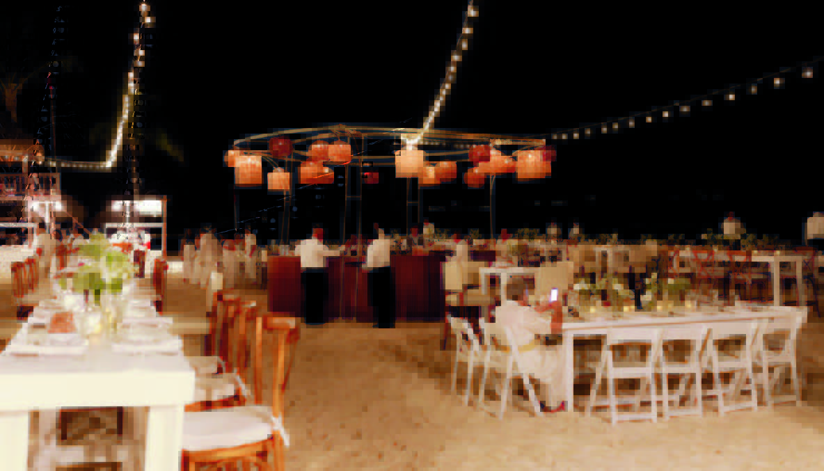 3 be all inclusive Night beach wedding reception with string lights, hanging lanterns, and long white tables set on the sand beneath a canopy of warm glow.