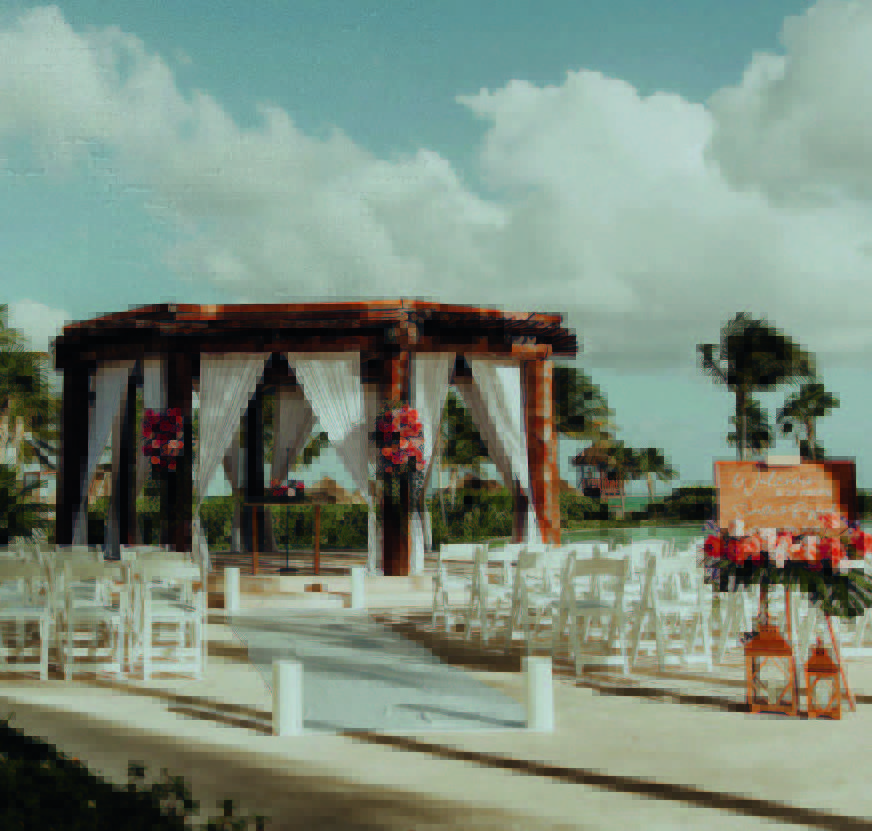 3 be all inclusive Beach wedding setup with a wooden gazebo, white drapes, and pink floral arrangements on a sandy aisle lined with white chairs.