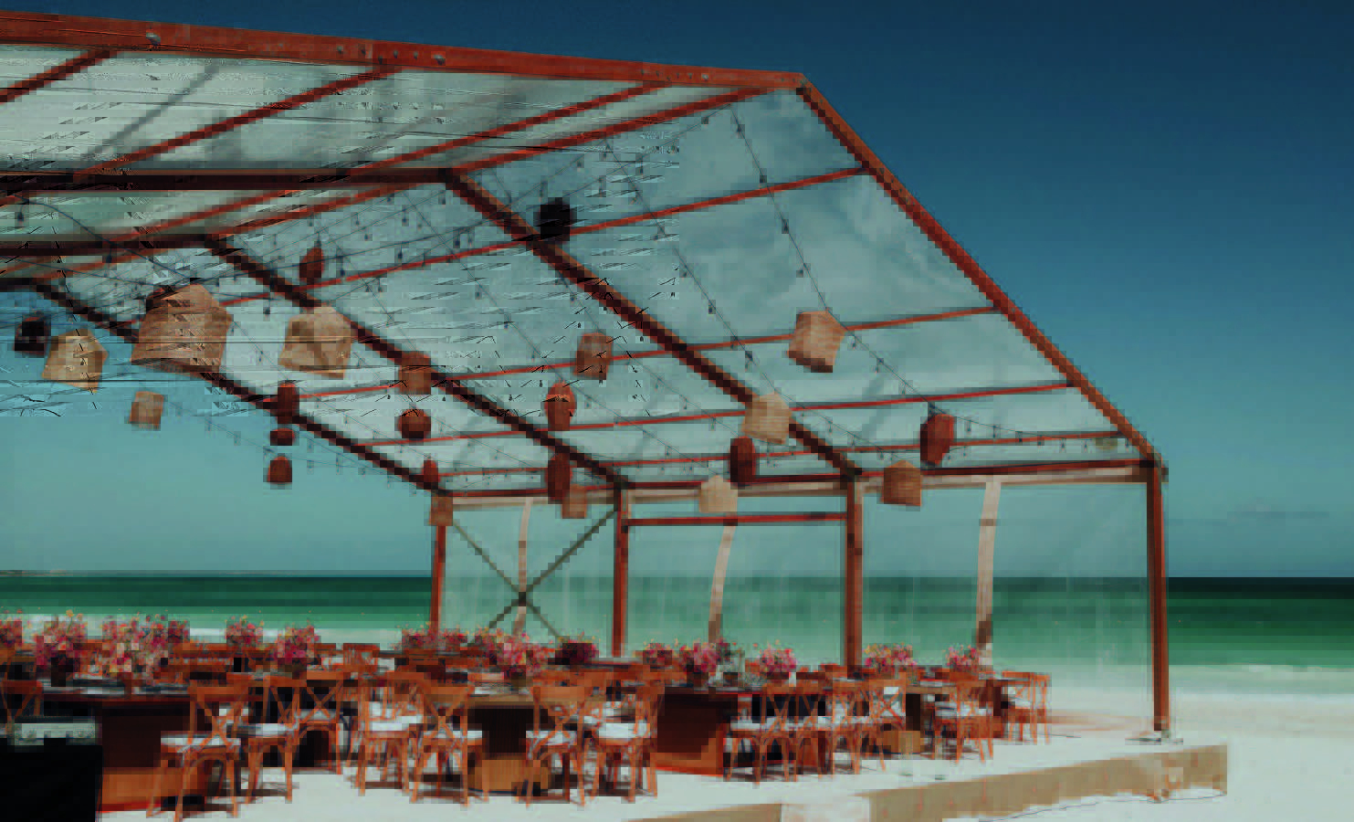 4 be all inclusive Beach wedding reception under a transparent canopy with string lights, lanterns, wooden chairs, and pink floral centerpieces on the sand by the ocean.