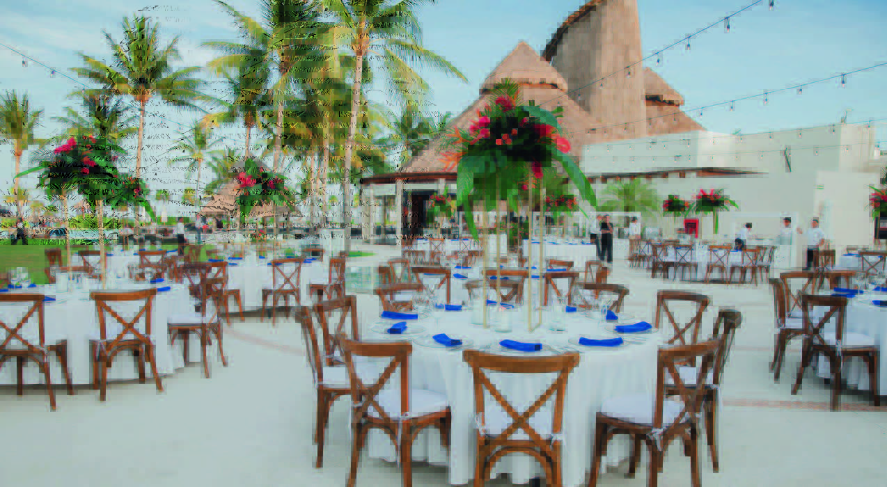 6 be all inclusive Outdoor tropical event setup with round tables, white cloths, blue napkins, tall floral centerpieces under palm trees and string lights at dusk.