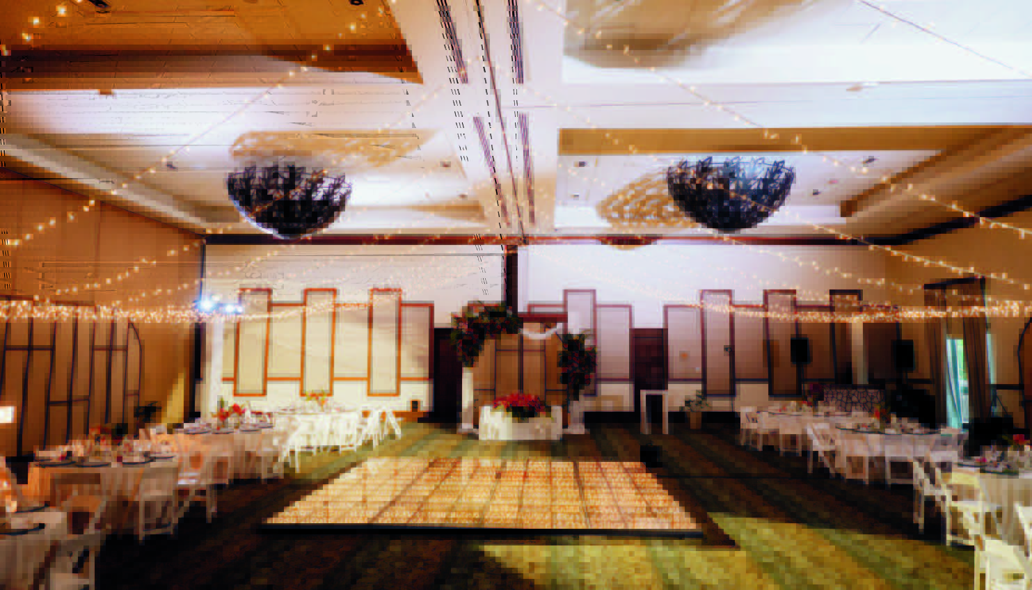 8 be all inclusive Banquet hall set for a celebration with round tables, white chairs, and string lights over a glowing square dance floor in the center.