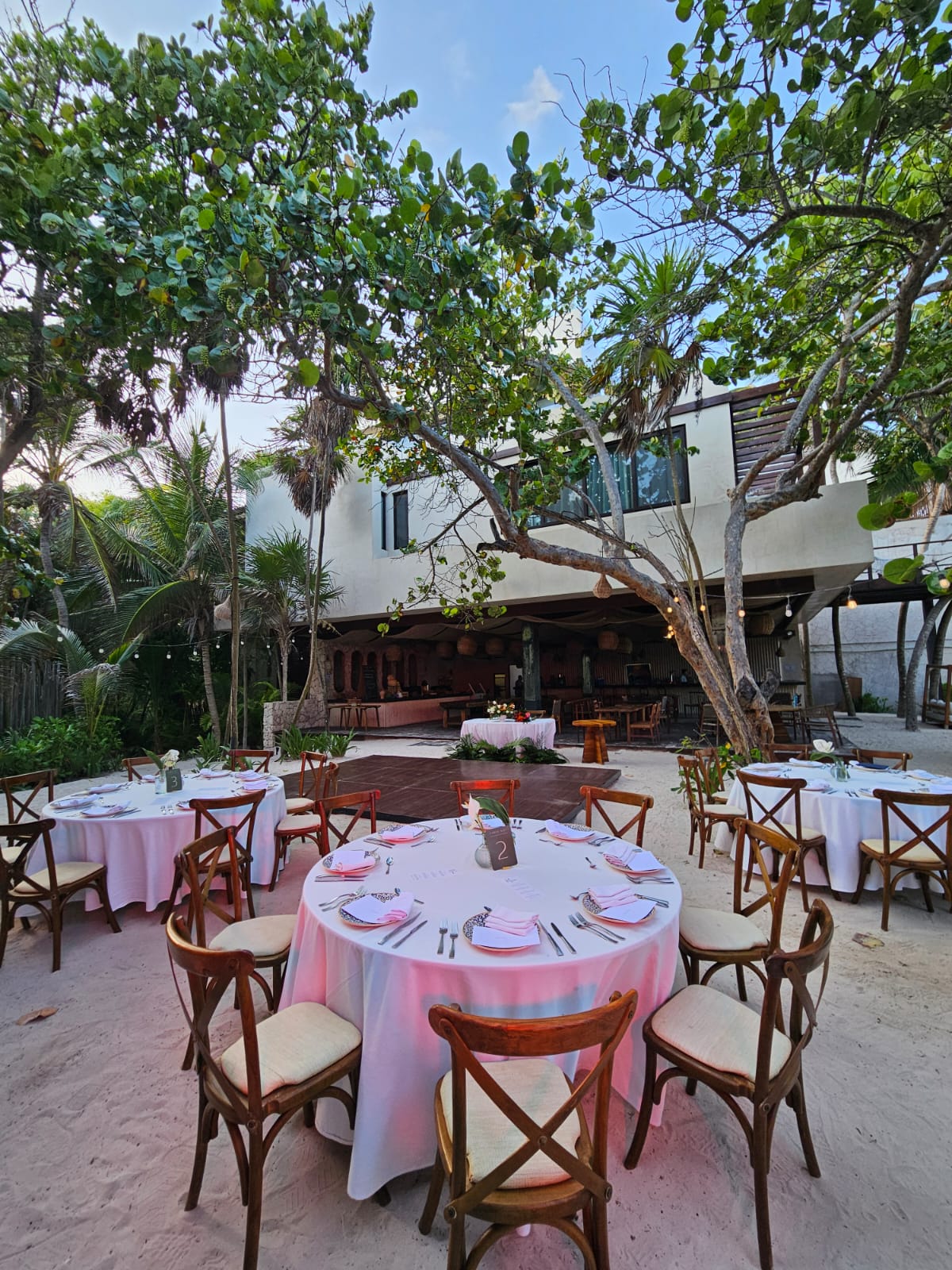 barracuda 2 be all inclusive Outdoor resort dining setup with pink-covered round tables and wooden chairs under leafy trees.