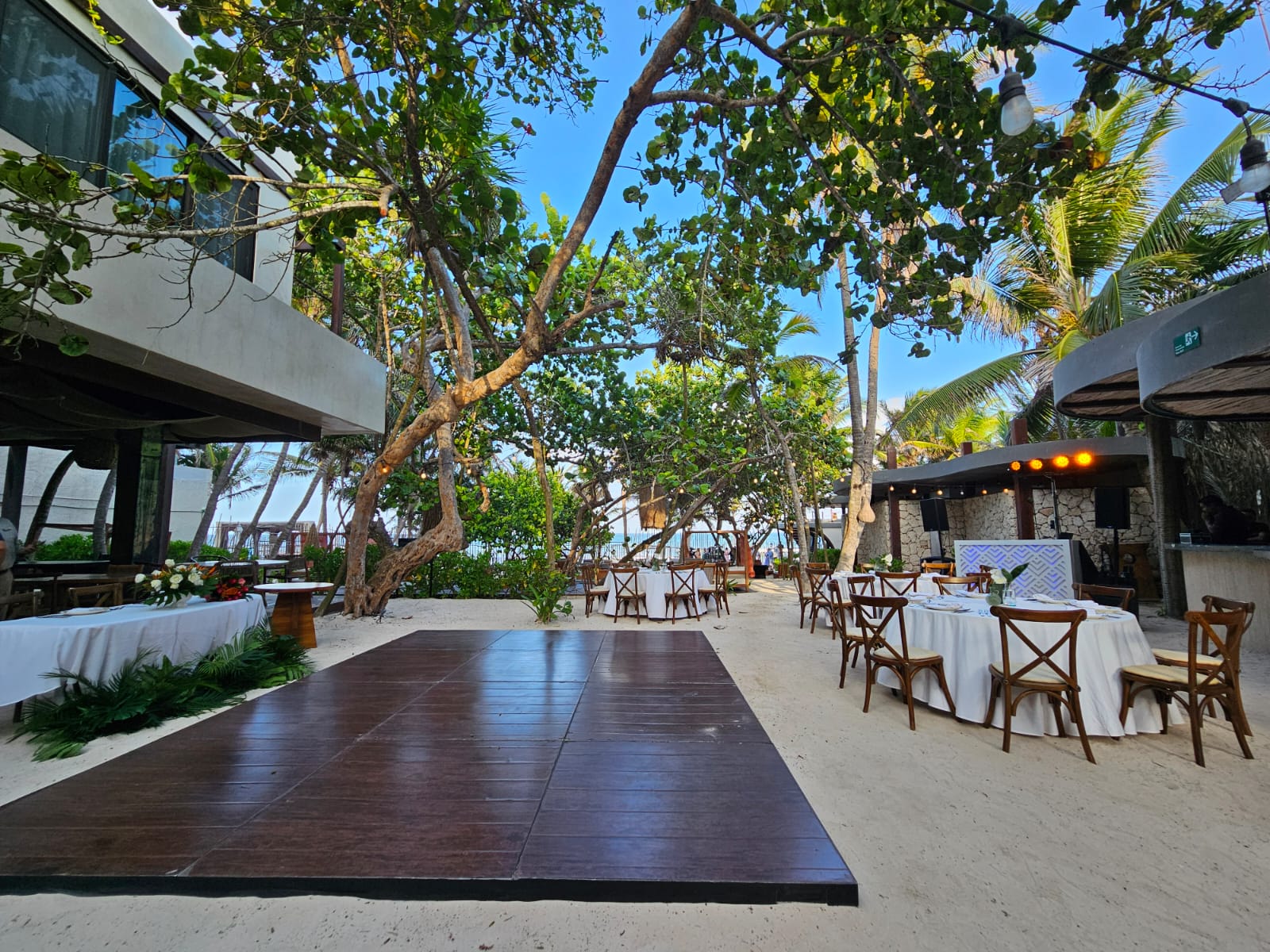 barracuda 5 be all inclusive Beachside event space with a wooden dance floor, round tables in white cloth, wooden chairs, and string lights among trees.