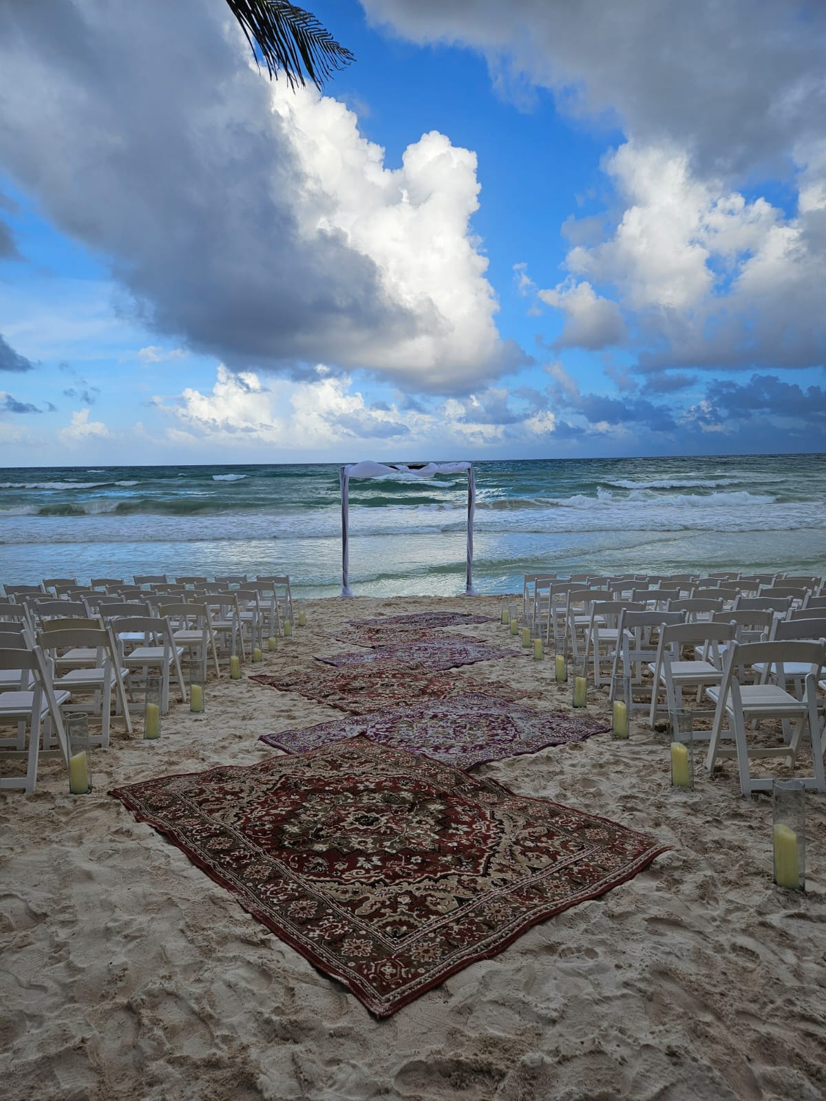 beach 1 be all inclusive Beach wedding setup on sand with white chairs lining an aisle, a burgundy carpet runner, and a white arch by the ocean under blue sky and clouds.