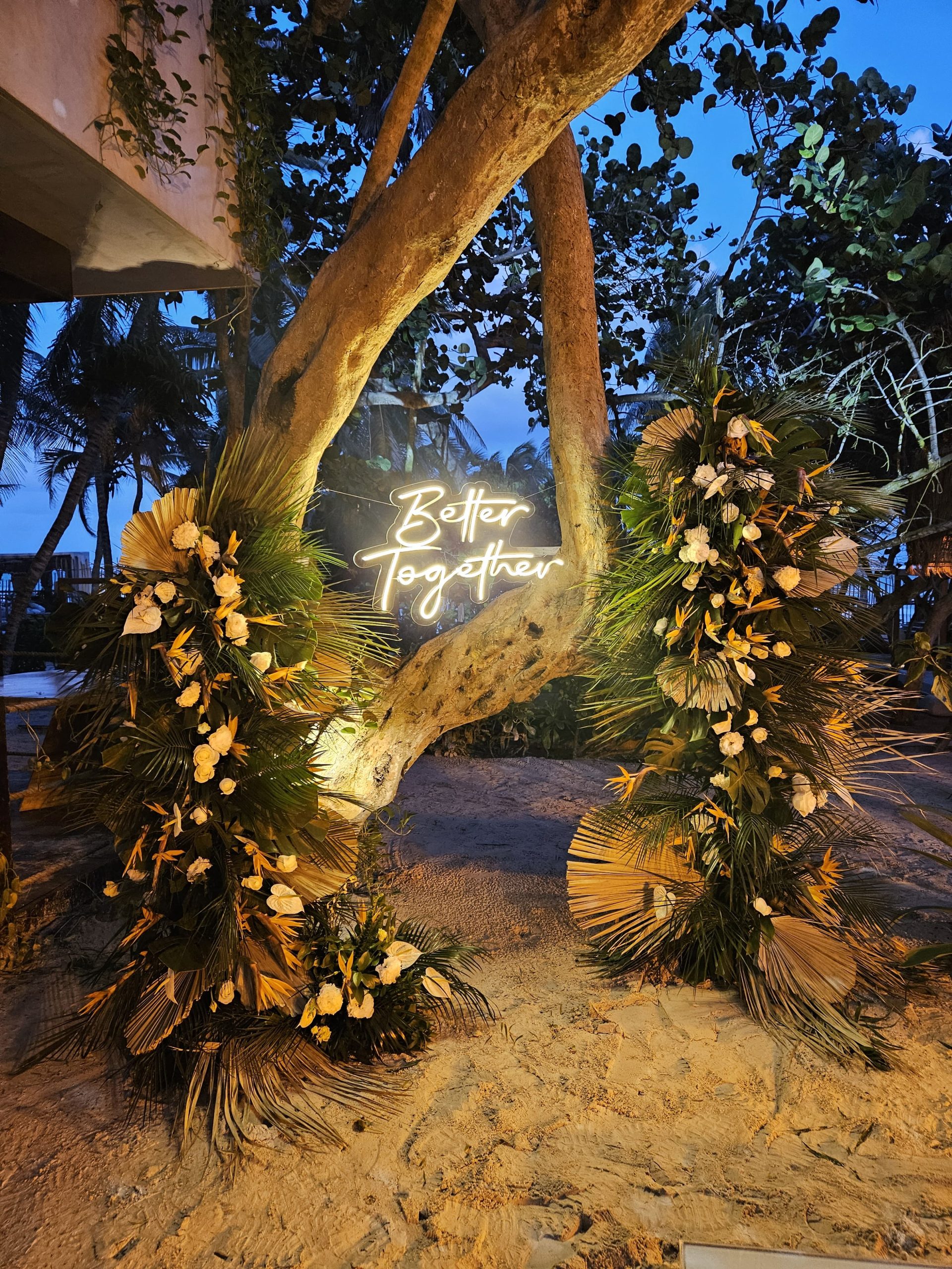 beach club 2 be all inclusive Tree wrapped in tropical floral garlands with a glowing neon sign reading 'Better Together' at dusk on a sandy beach setting