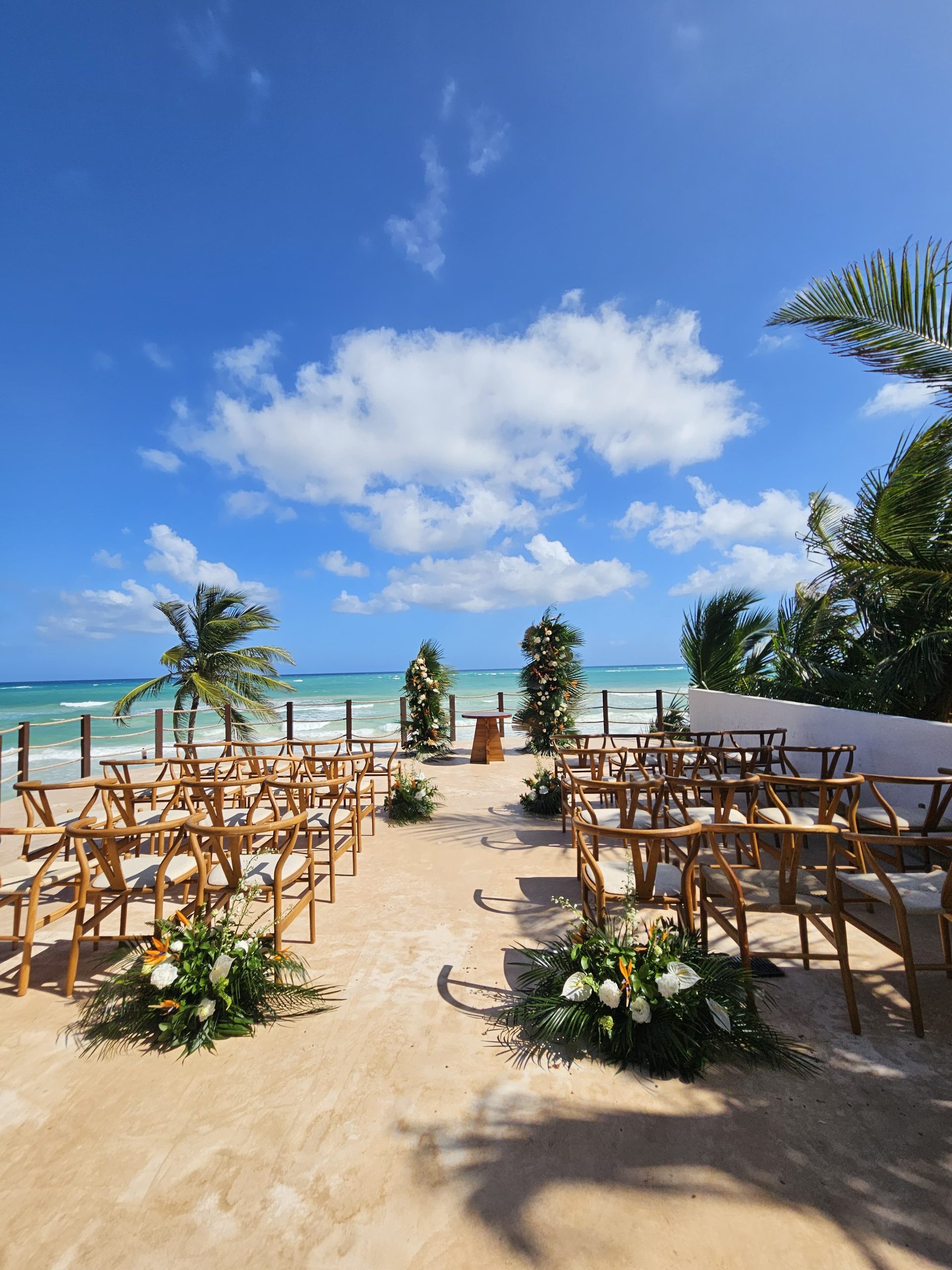 beach club 4 be all inclusive Beachside wedding setup with wooden chairs facing the ocean and floral arches along a sandy aisle under a blue sky.