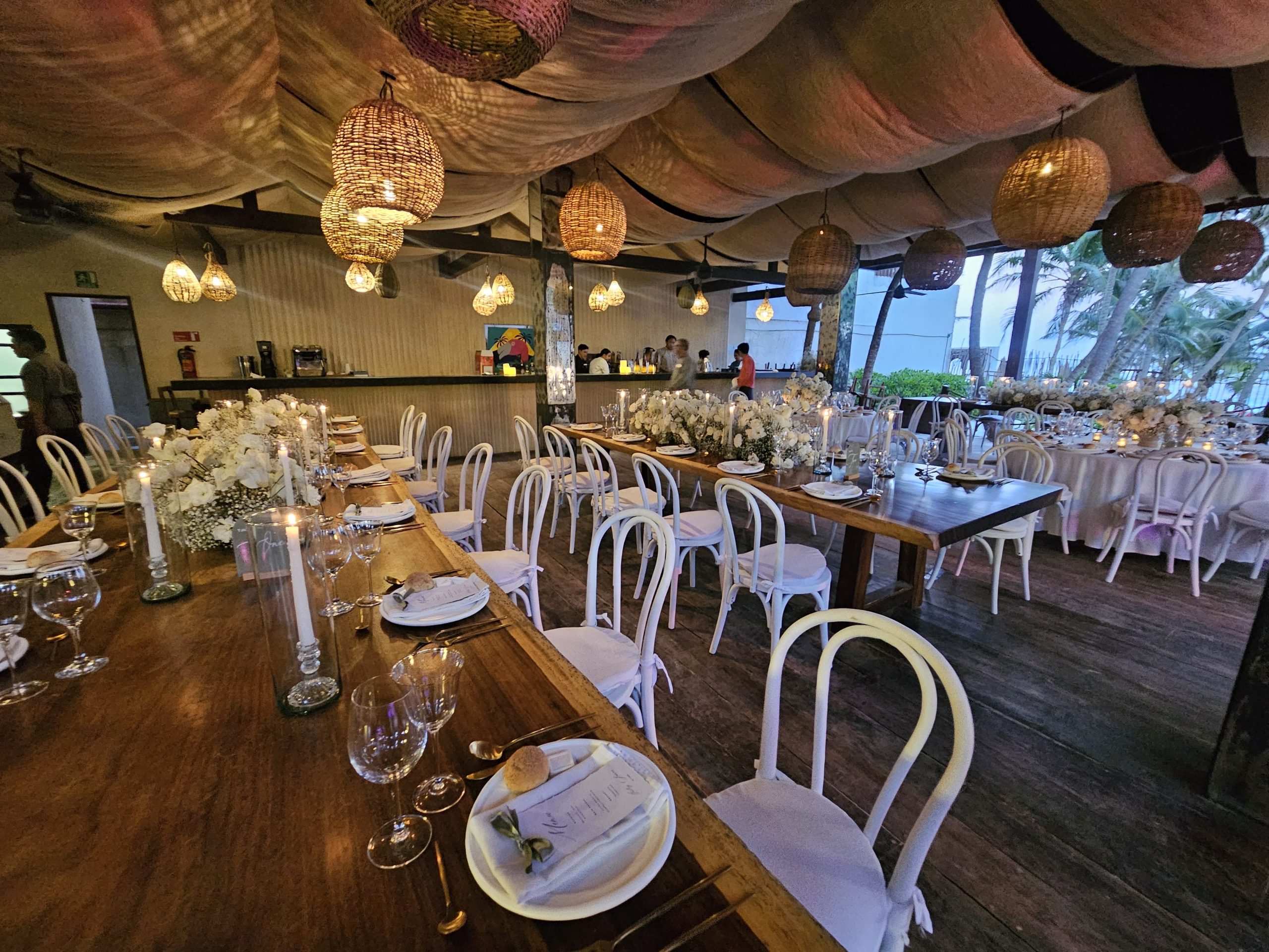 beach club 7 be all inclusive Long wooden banquet tables with white chairs, floral centerpieces, candles, and woven pendant lamps in a tropical wedding venue at dusk.