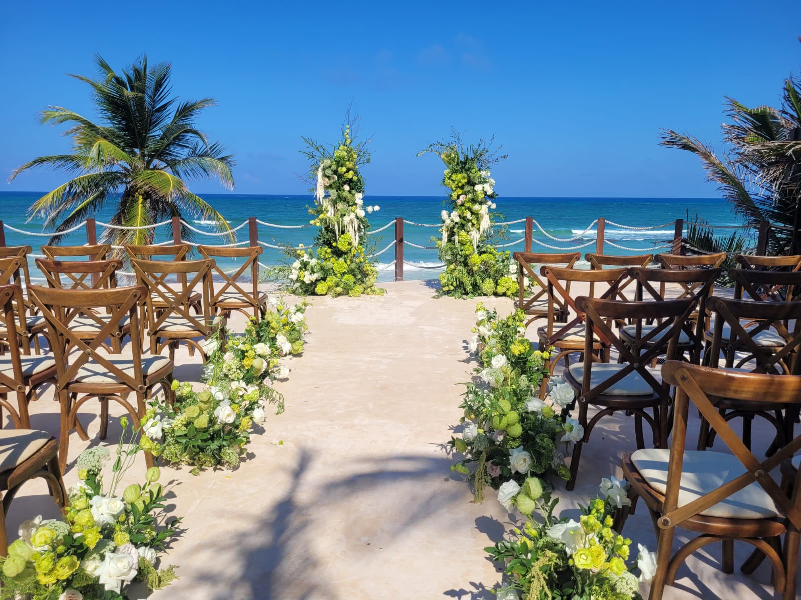 beach club 9 be all inclusive Beach wedding setup: wooden chairs facing a central aisle lined with green‑and‑white floral arrangements, two floral arches, and the ocean in the background.