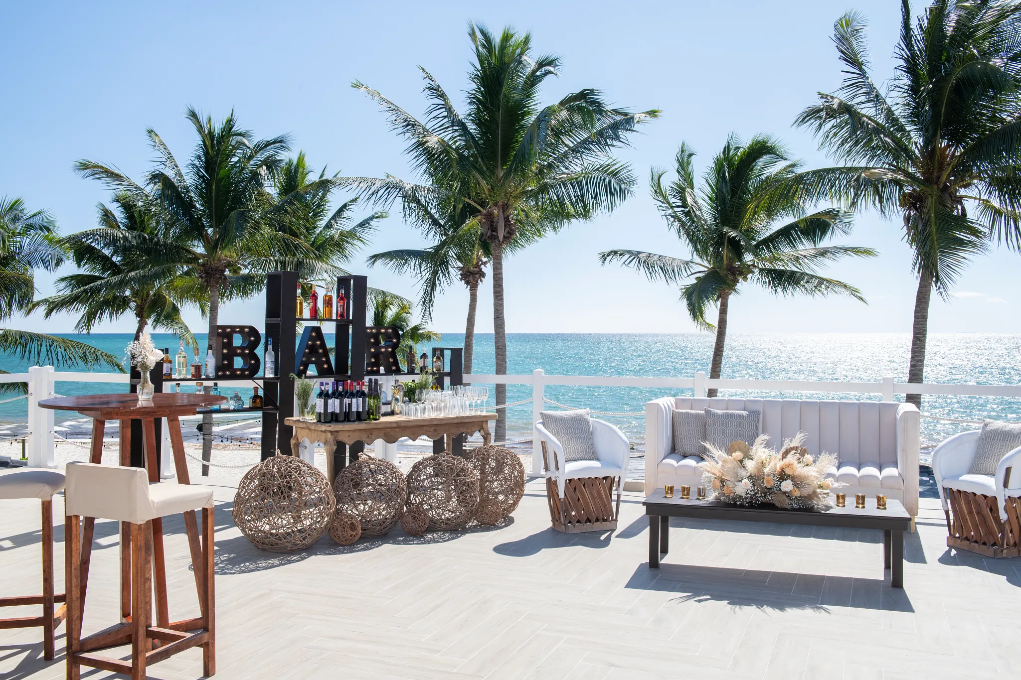 caniap0051micelolarooftoploungesetup be all inclusive Beachside bar setup with bottles, glasses, and seating against a ocean sunrise, palm trees framing the scene