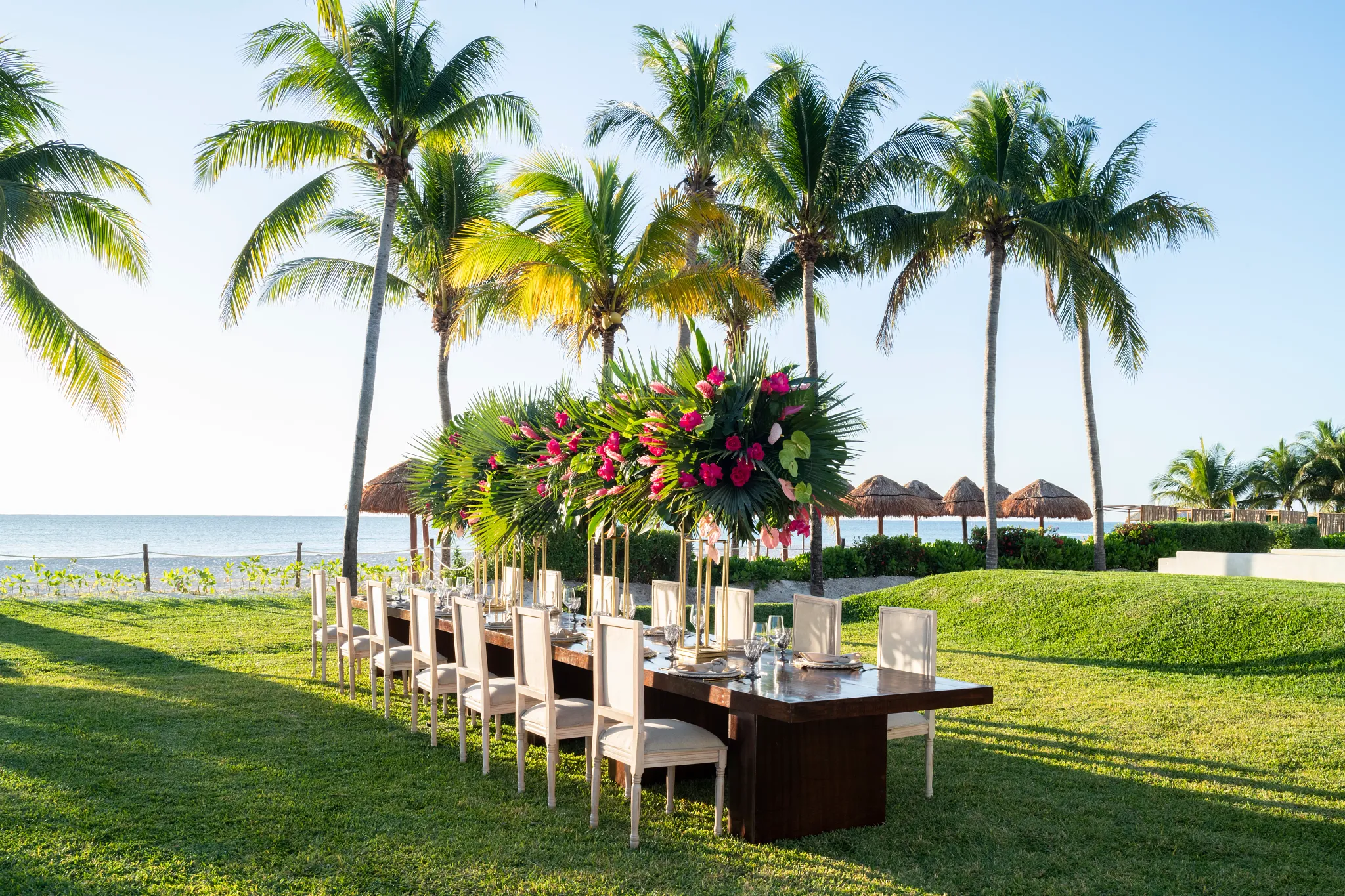 caniap0081gardenweddingreception be all inclusive Long outdoor beachside dining table set on green lawn, with palm trees and ocean in the background, and a tropical floral centerpiece.