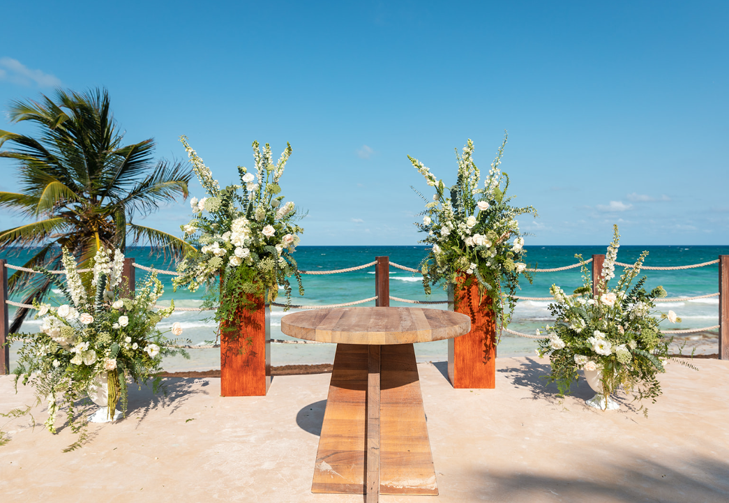 ceremonia 1 be all inclusive Beachside wedding altar setup with three tall white-flowered arrangements in wooden vases and a circular wooden table, against blue ocean and clear sky.