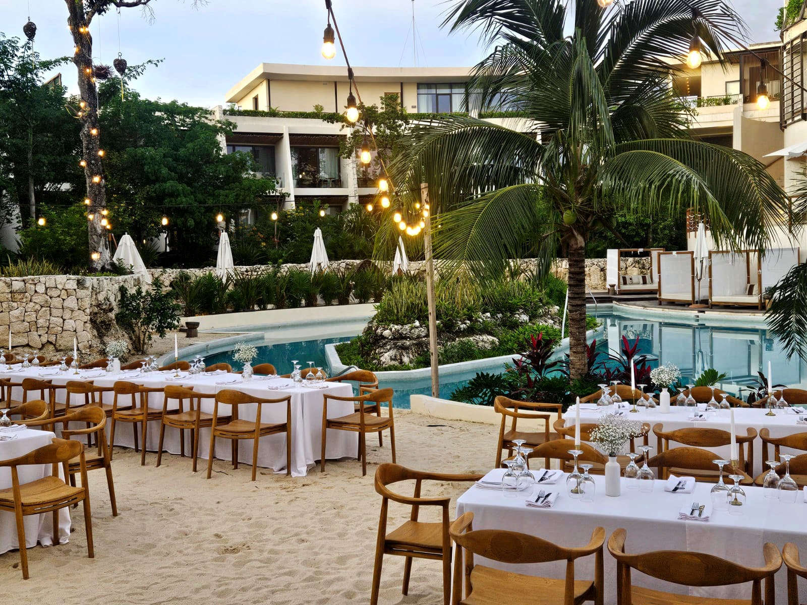 cenote central 3 be all inclusive Outdoor beachside dinner setup with long white-tablecloth tables, wooden chairs, candles, and string lights by a shallow pool.