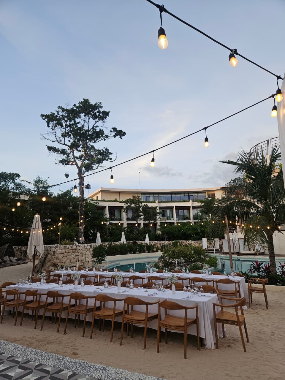 cenote central 7 be all inclusive Outdoor beachside banquet setup with a long white-clothed table, wooden chairs, string lights, and a pool with a modern building in the background, at dusk.