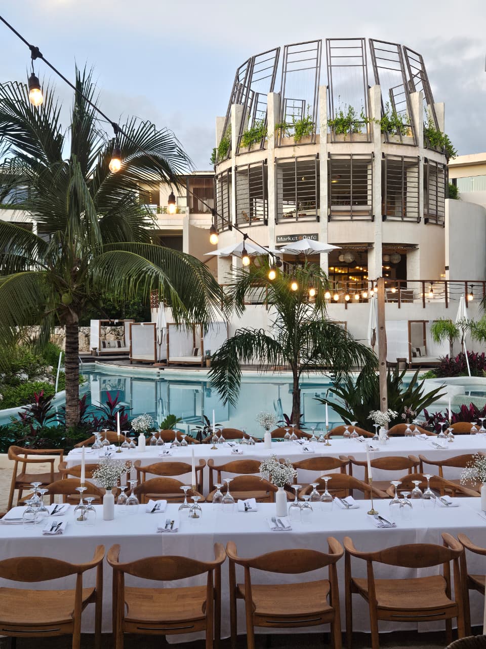cenote central 8 be all inclusive Poolside dining setup with long white tables, wooden chairs, glassware, candles, and tropical plants under string lights.