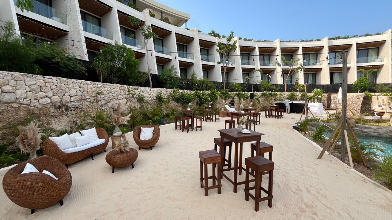 cenote luna 4 be all inclusive Beachside resort lounge area on sand with wicker chairs and sofas, white cushions, and wooden high tables, stone retaining wall, string lights, and a pool to the right.