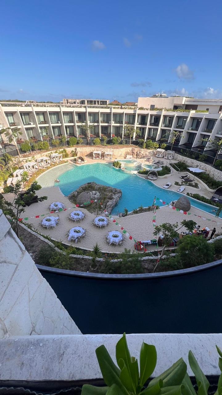 cenote luna 5 be all inclusive Resort pool area with a winding turquoise pool, surrounding white-walled buildings, rock features, and round tables with chairs under umbrellas.