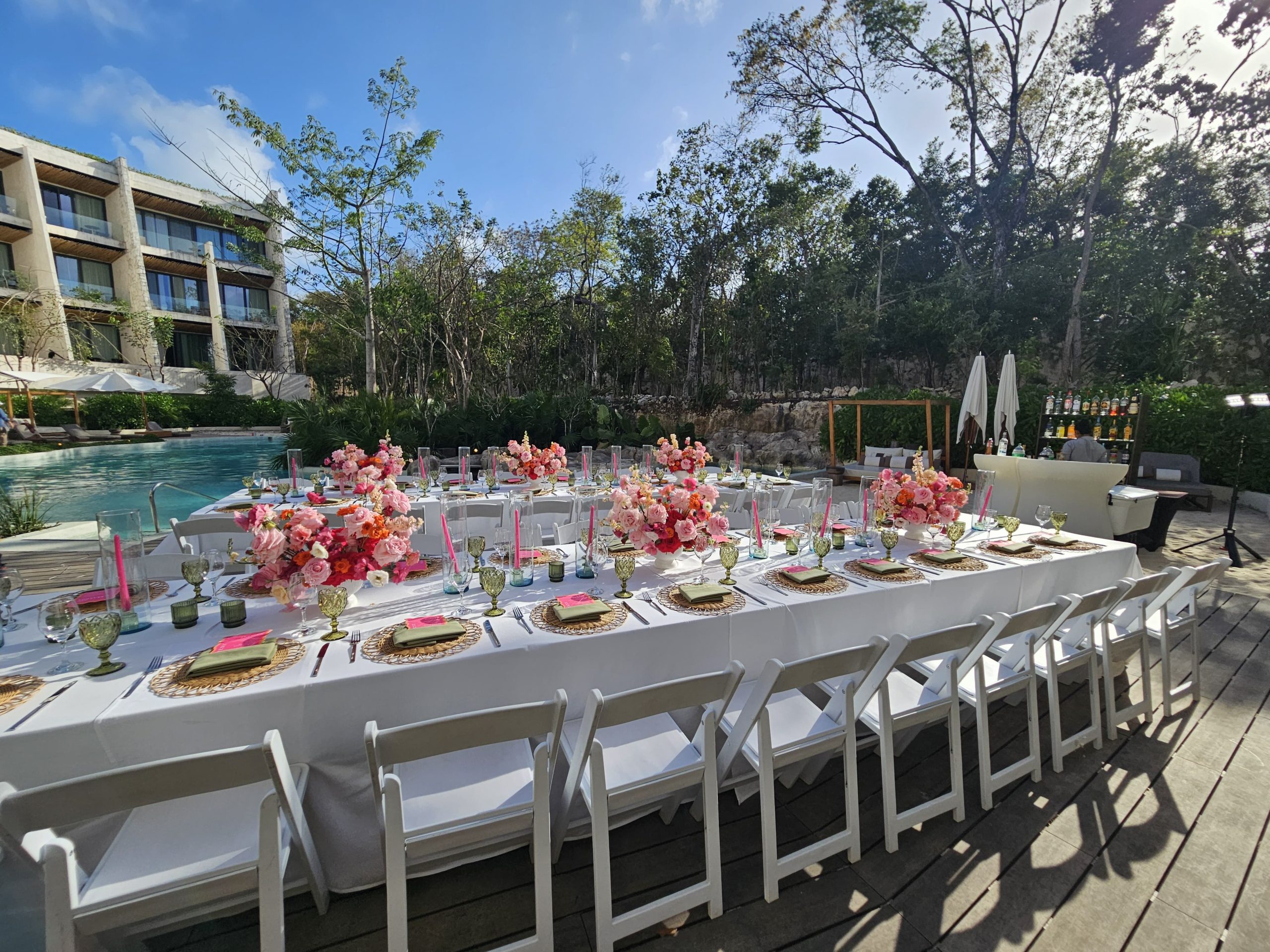 cenote zen 3 be all inclusive Poolside banquet setup with long white tables, pink flower centerpieces, glassware, and pink candles by a hotel building.