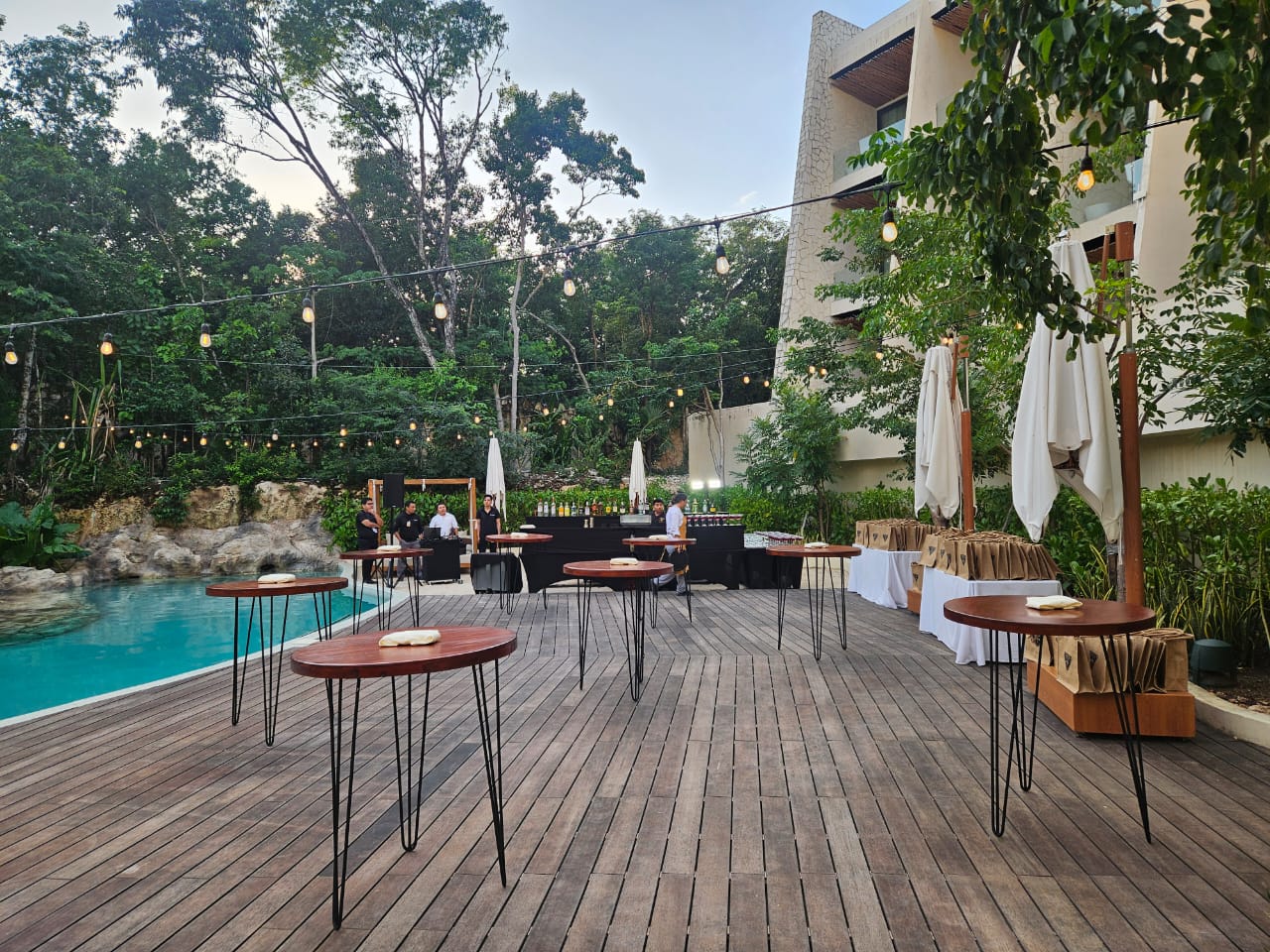 cenote zen 5 be all inclusive Outdoor poolside event with high wooden tables on a deck and string lights above, greenery in the background, and a bar area in the distance.","A courtyard setup for a reception: round high tables, warm string lights, and a catering bar along the back with staff milling about.","Sunlit poolside lounge with cocktail tables and a staffed bar, surrounded by trees and a modern building.","Evening-style outdoor party setup by a pool: tall tables, umbrellas, and a long beverage station under string lights.","Casual outdoor cocktail area by a pool, featuring wooden-topped tables on hairpin legs and a bar service area in the rear."