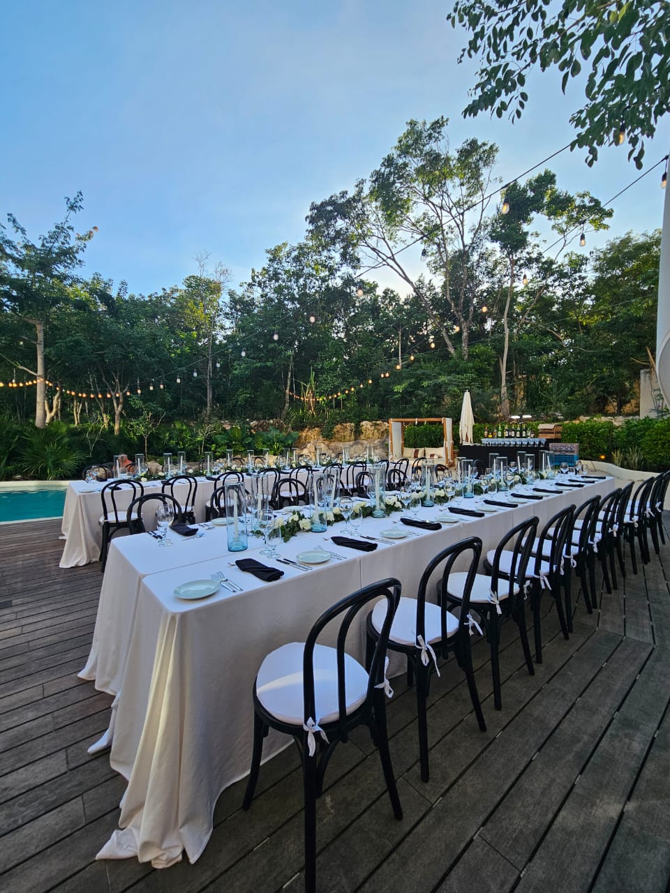 cenote zen 6 be all inclusive Outdoor formal dining setup with long rectangular tables, white tablecloths, black chairs, and string lights among trees.