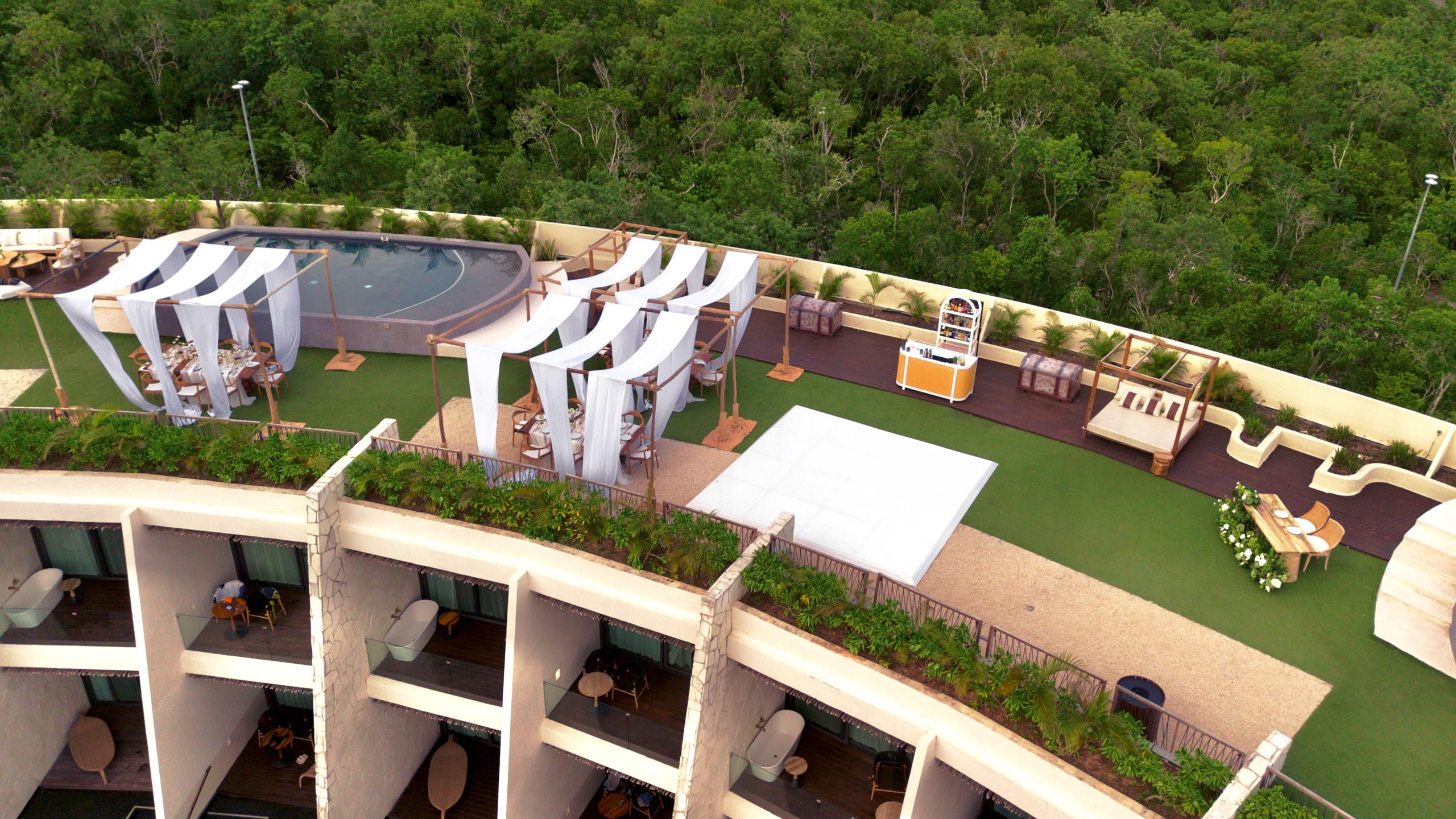 dji0726 be all inclusive Aerial view of a rooftop resort terrace with white fabric cabanas, a circular pool, and lounge seating surrounded by greenery.