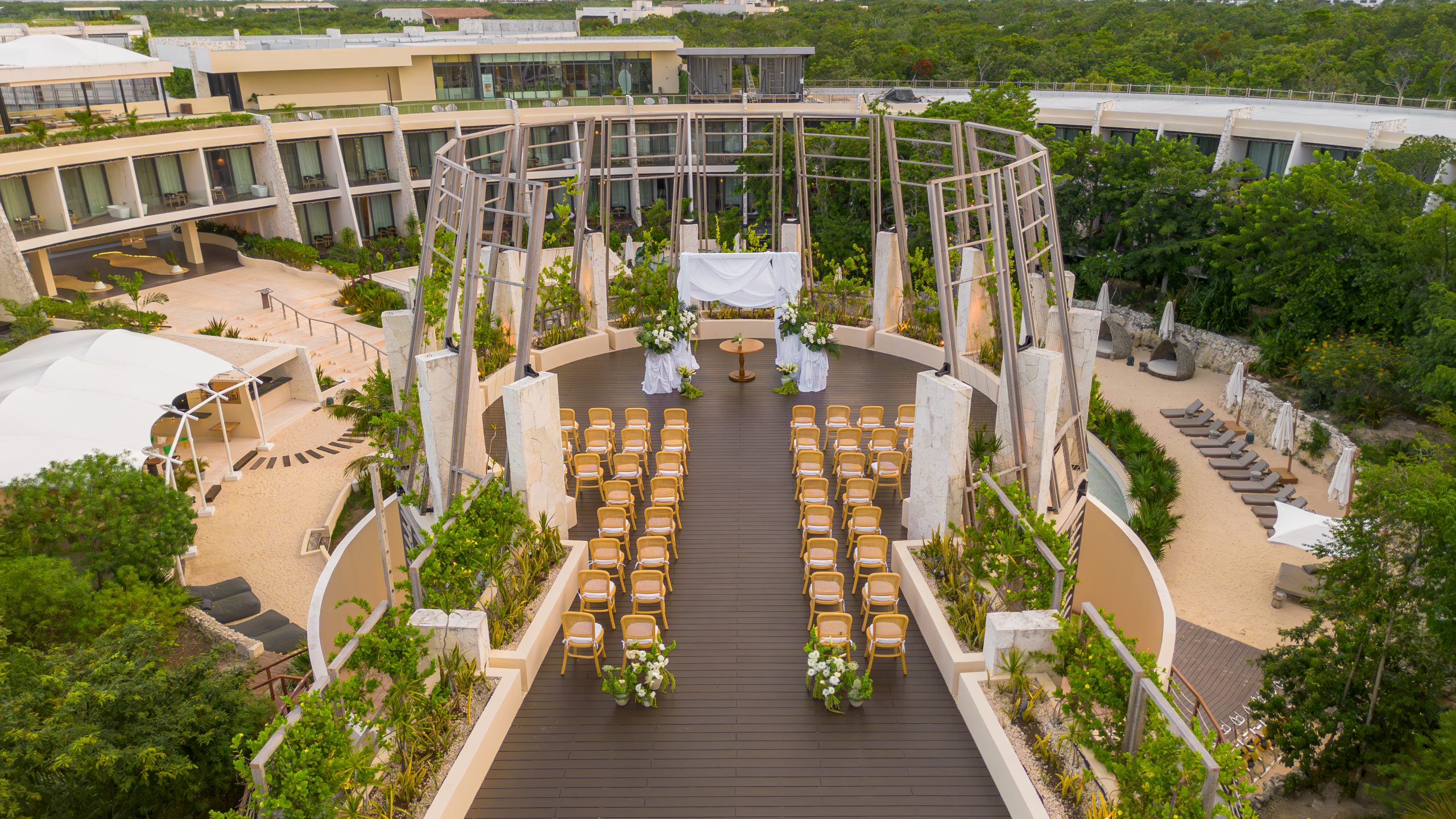 dji0744 be all inclusive Outdoor wedding setup on a wooden deck with rows of cream chairs facing a draped altar and floral arrangements.