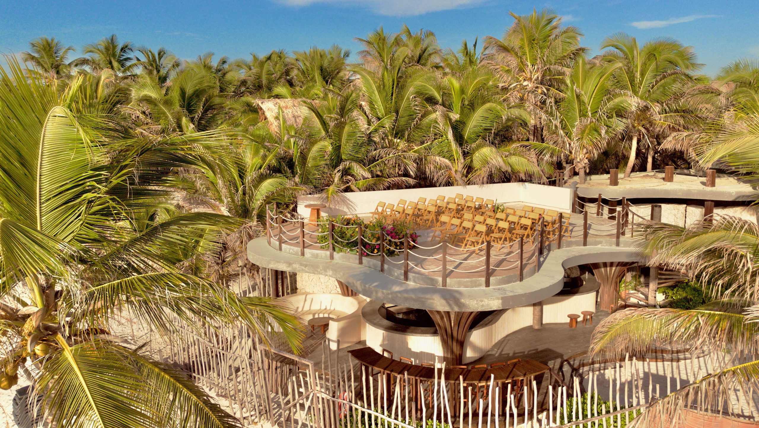 dji0770 be all inclusive Circular, elevated seating platform with rows of beige wooden chairs, surrounded by dense palm trees under a bright blue sky.