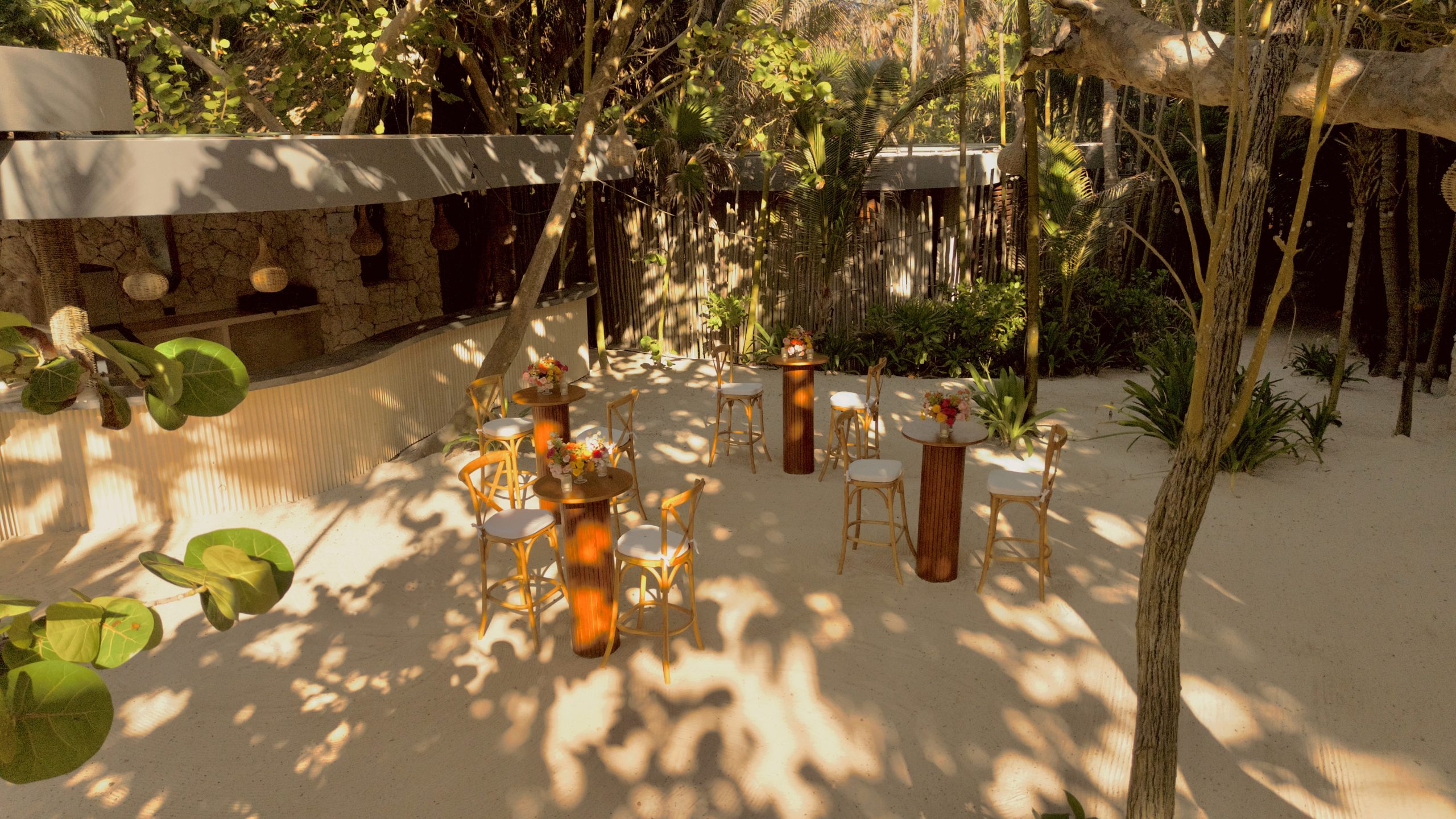 dji0796 be all inclusive Outdoor tropical event setup on sand with round tables, tall wooden cocktail tables, and white-stool seating surrounded by lush trees and hanging lanterns.