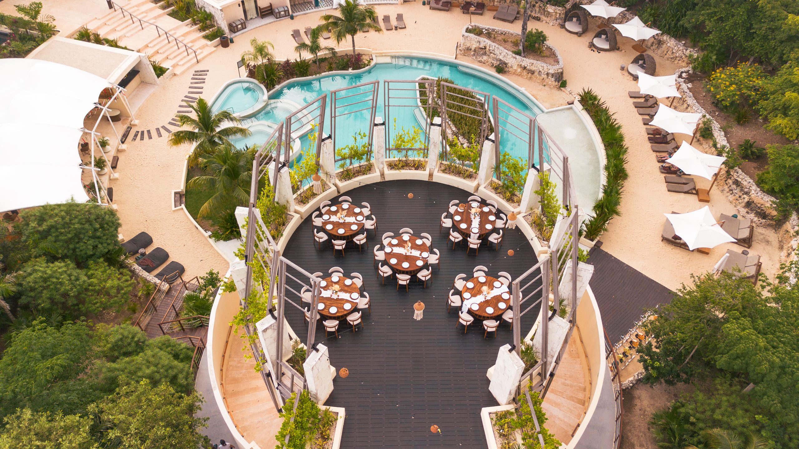dji0942 be all inclusive Aerial view of a resort pool and circular dining terrace with round tables set for guests, surrounded by palm trees and sun loungers.