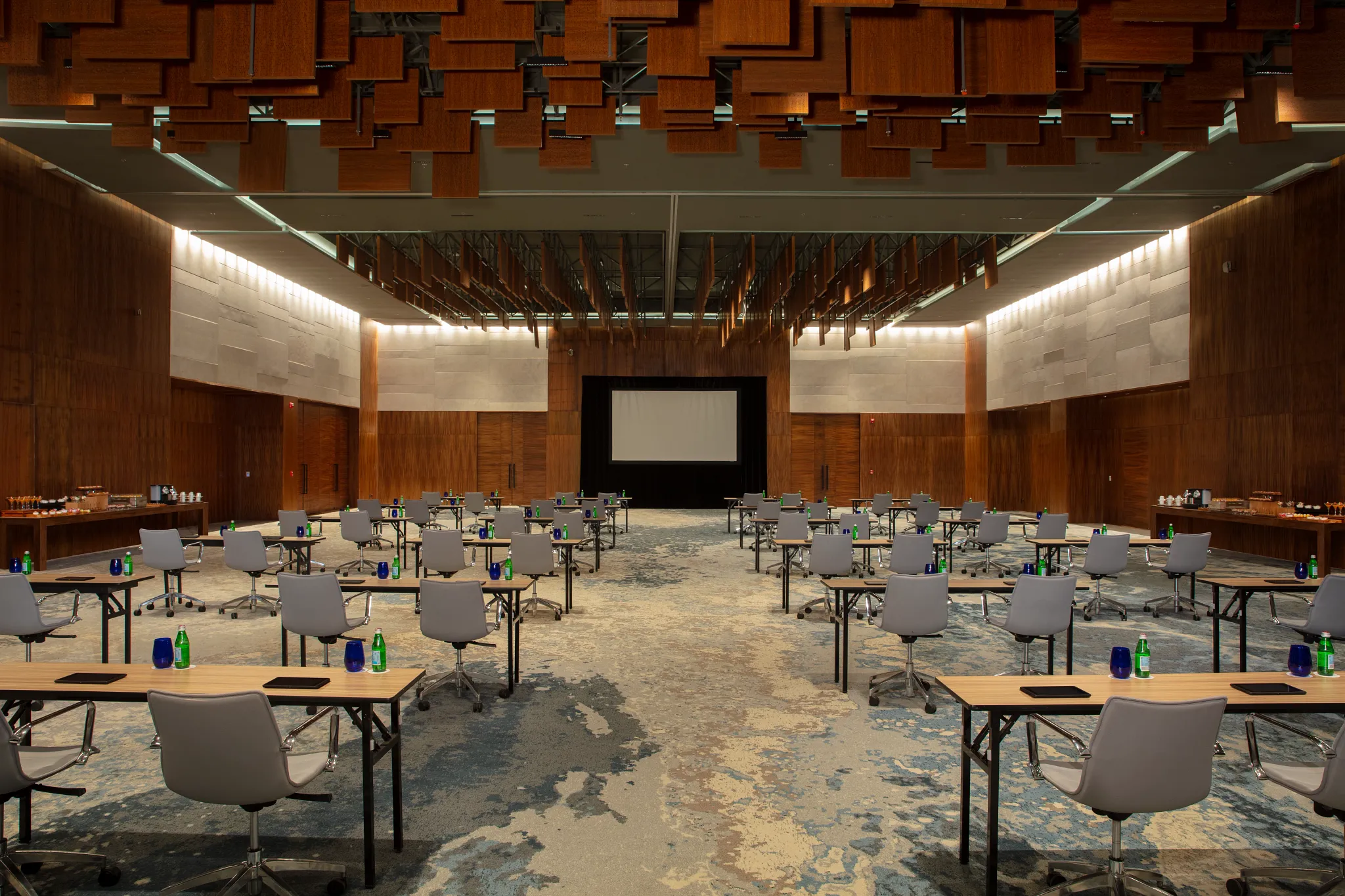 drbmigrpballroom1acb be all inclusive Large conference room set up with rows of tables and gray chairs facing a projector screen at the far end; bottles and notebooks on tables.