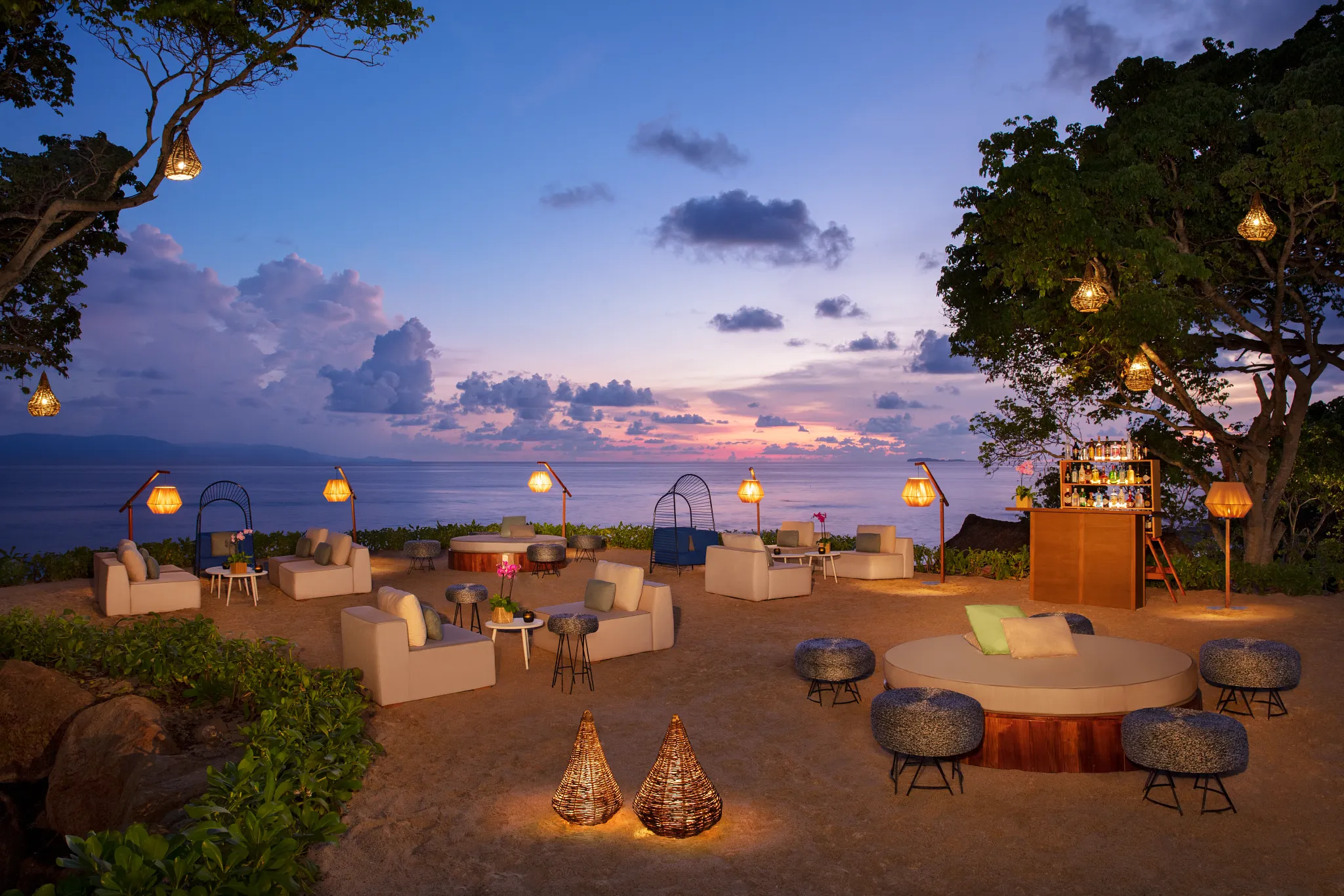drbmigrpbeachcocktail3acb be all inclusive Beachside lounge at dusk with glowing woven lanterns, white sofas, and a bar overlooking the ocean.